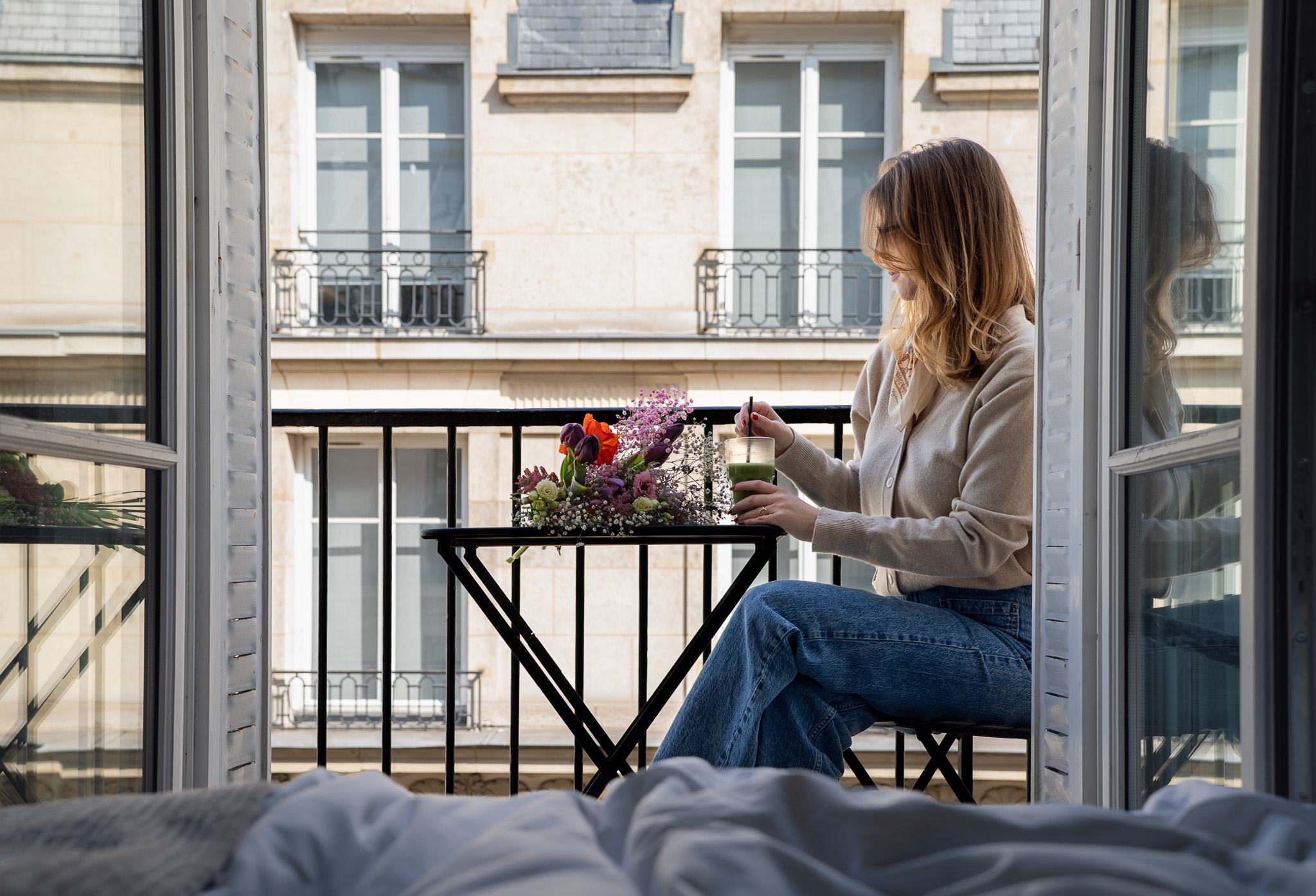 Une femme aux cheveux clairs porte un gilet crème et un jean bleu, assise sur un petit balcon parisien avec une table noire pliable devant elle décorée d’un bouquet de fleurs colorées, tenant un verre avec une boisson verte et une paille, en regardant vers la droite, entourée par des fenêtres et des volets blancs ouverts.