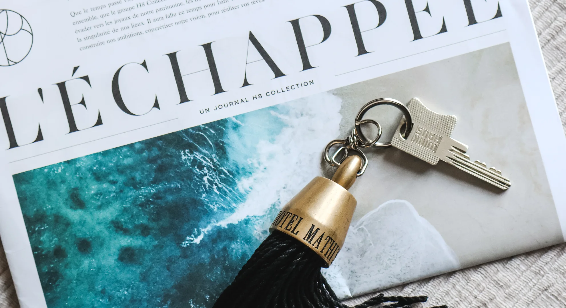 Une clé d'hôtel argentée attachée à un porte-clés à pompon noir et or portant l'inscription « HOTEL MATHIS », posée sur un journal avec une grande photo d'océan et le titre partiel « L'ÉCHAPPEE » en lettres noires.