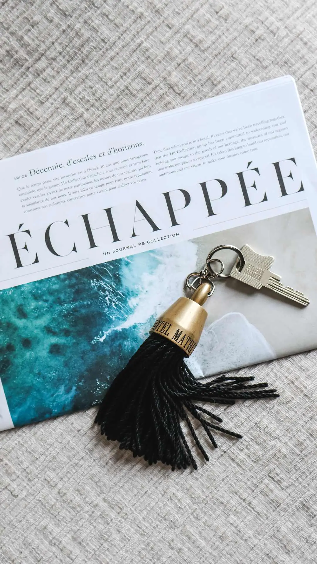 Une clé avec un porte-clés en forme de gland noir et doré marqué Hôtel Mathis posé sur un magazine Échappée montrant une photo de l'océan.