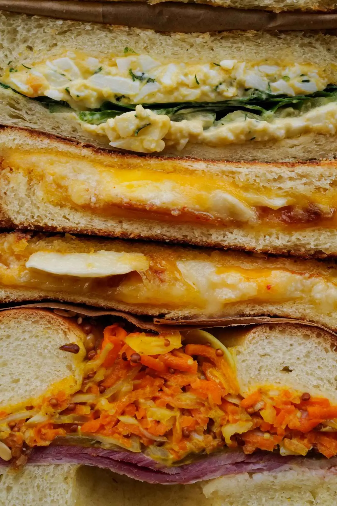 Trois sandwichs empilés en gros plan : un sandwich aux œufs avec des morceaux d'œufs durs et de la laitue, un sandwich au fromage fondu, et un sandwich garni de carottes râpées, cornichons et jambon sur du pain en tranches.