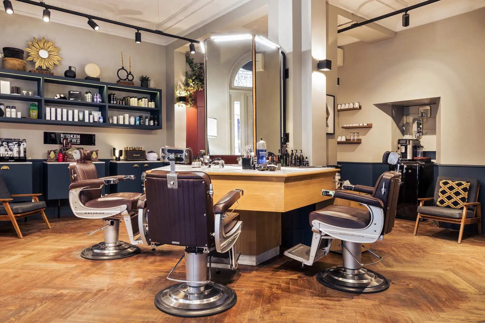 Salon de coiffure moderne avec trois fauteuils de barbier en cuir marron autour d'un îlot en bois avec miroir, étagères avec produits capillaires et décorations murales, sol en parquet.