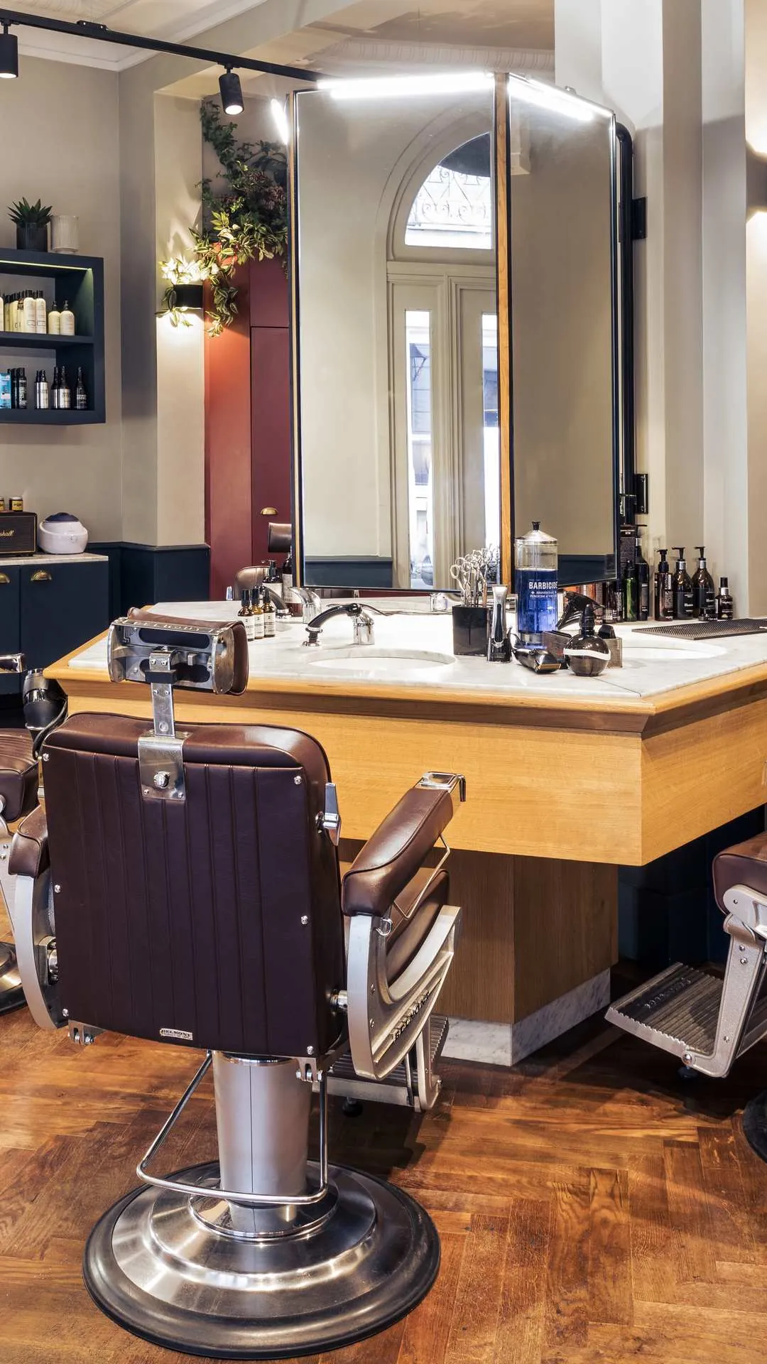 Chaise de barbier en cuir marron devant un grand miroir éclairé avec lavabo et divers produits de coiffure sur le comptoir en bois dans un salon de coiffure élégant avec sol en bois et murs clairs.