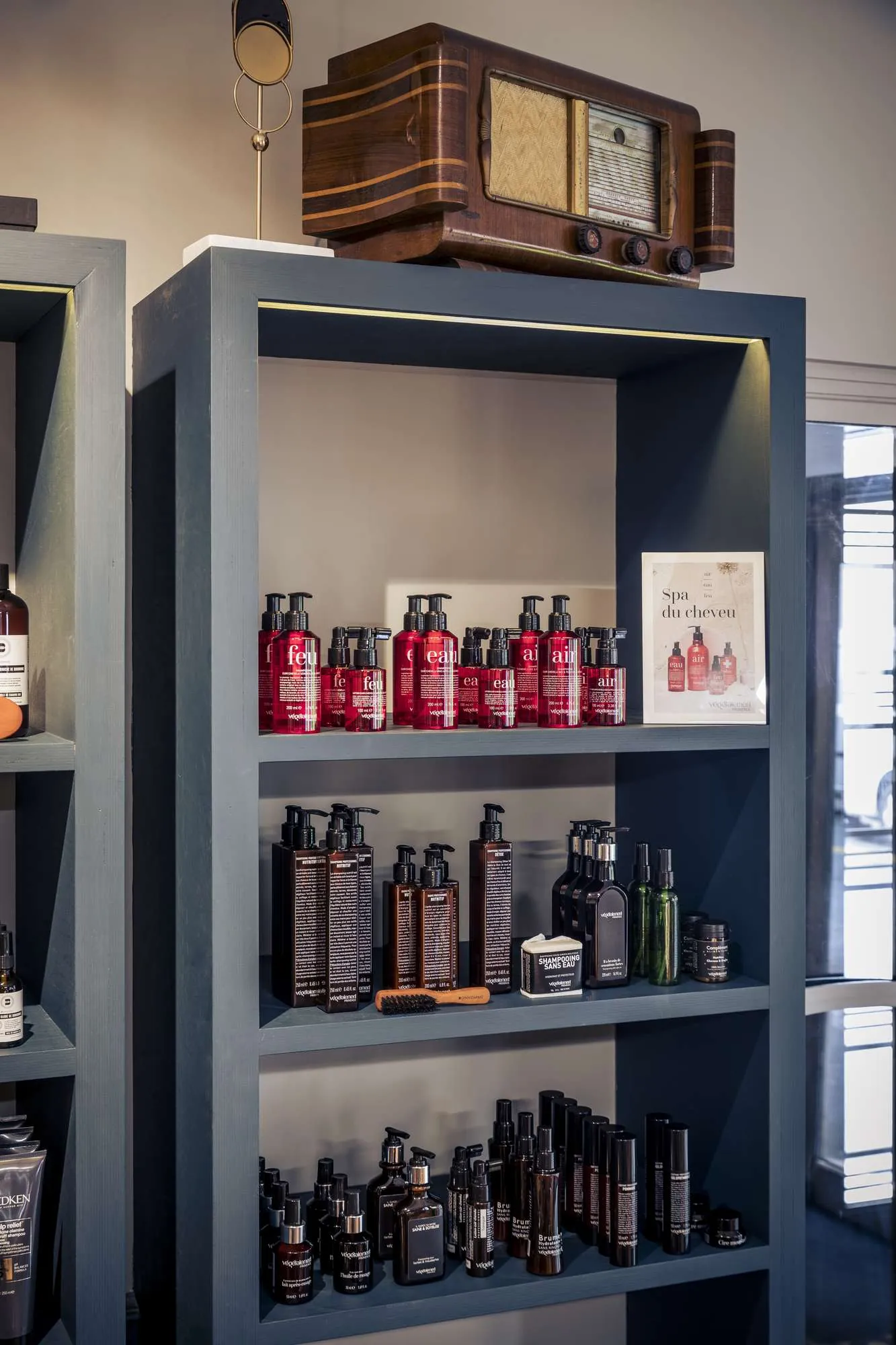 Étagère grise contenant plusieurs produits de soins capillaires en bouteilles rouges, noires et vertes, une brosse en bois et un petit panneau indiquant "Spa du cheveu". Un vieux poste de radio en bois est posé sur le dessus de l'étagère.
