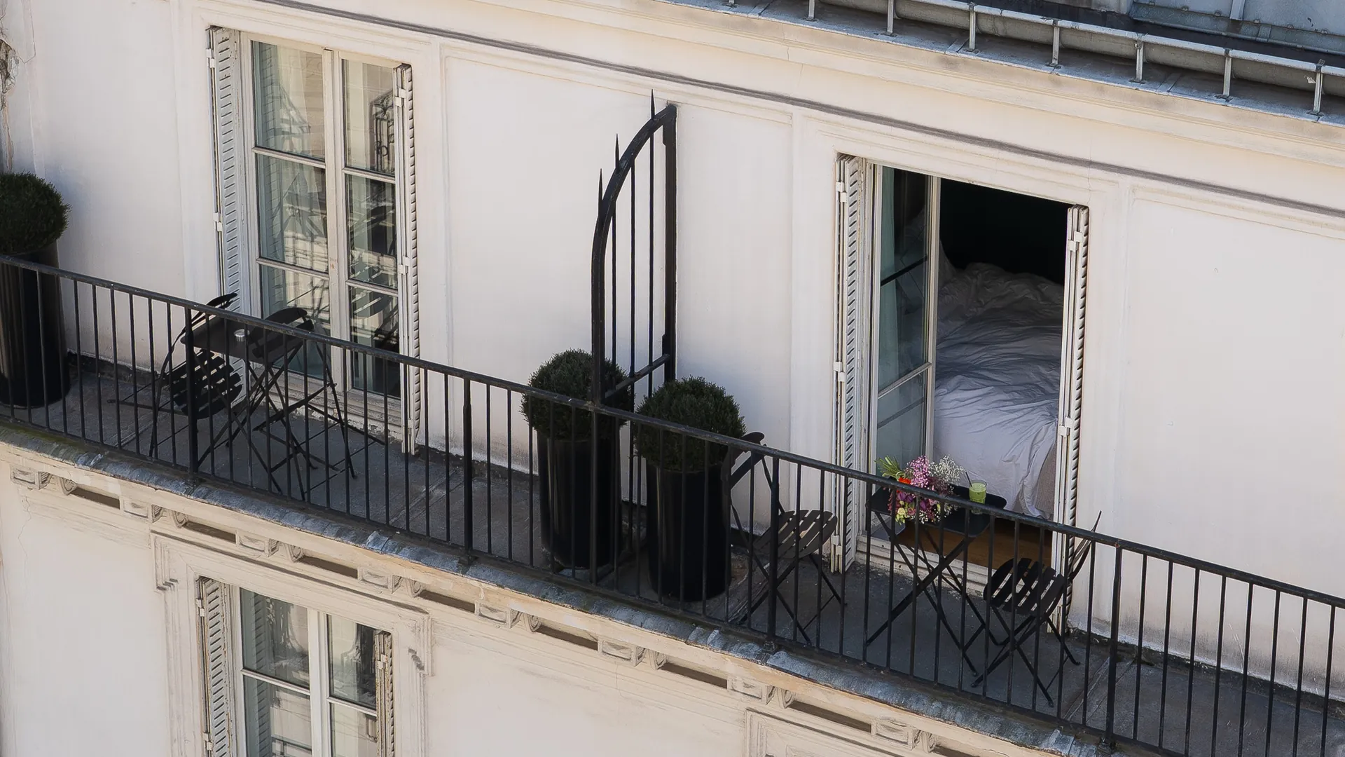 Balcon d'un immeuble avec une rampe en métal noir, deux petites tables avec chaises pliantes, deux grands pots noirs avec des arbustes, une porte fenêtre entrouverte donnant sur une chambre avec un lit défait et un bouquet de fleurs sur la rambarde.
