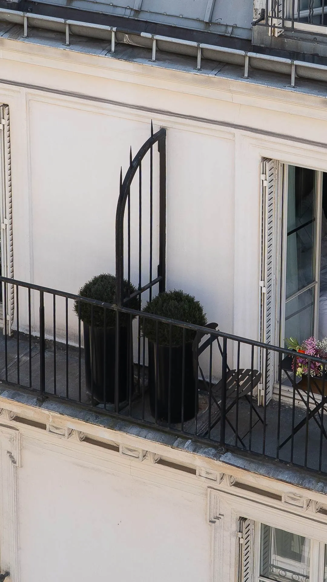 Balcon en fer forgé avec deux grands pots noirs contenant des buissons taillés, une chaise pliante noire, une table avec un bouquet de fleurs colorées, et une porte-fenêtre ouverte avec volets blancs sur un mur crème.
