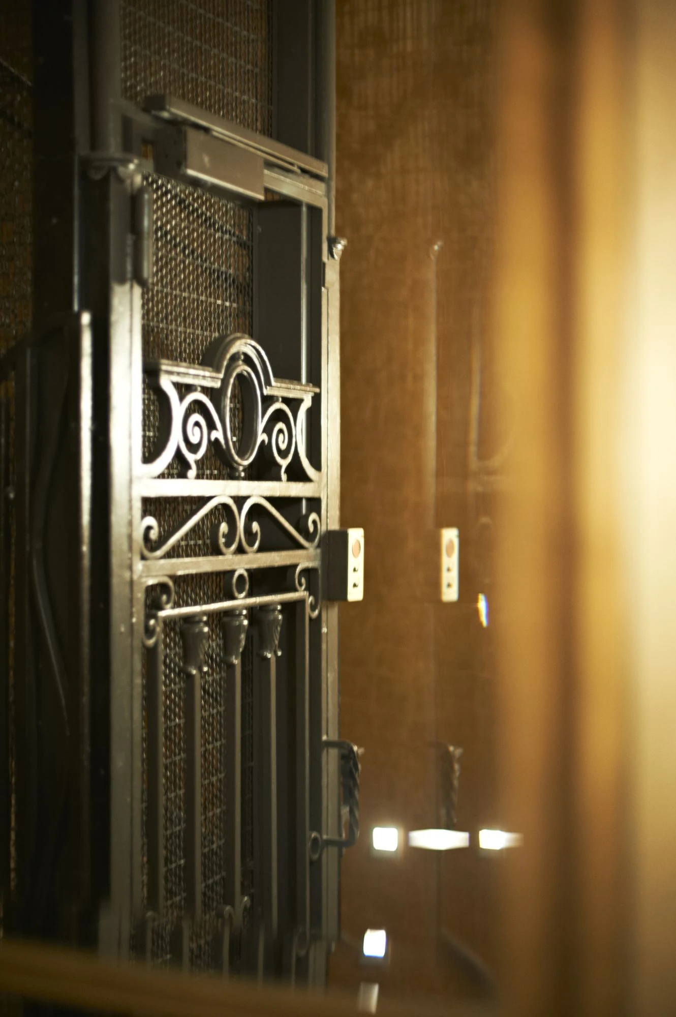 Porte d'ascenseur ancienne en métal noir avec motifs décoratifs et bouton d'appel, éclairée par une lumière dorée.