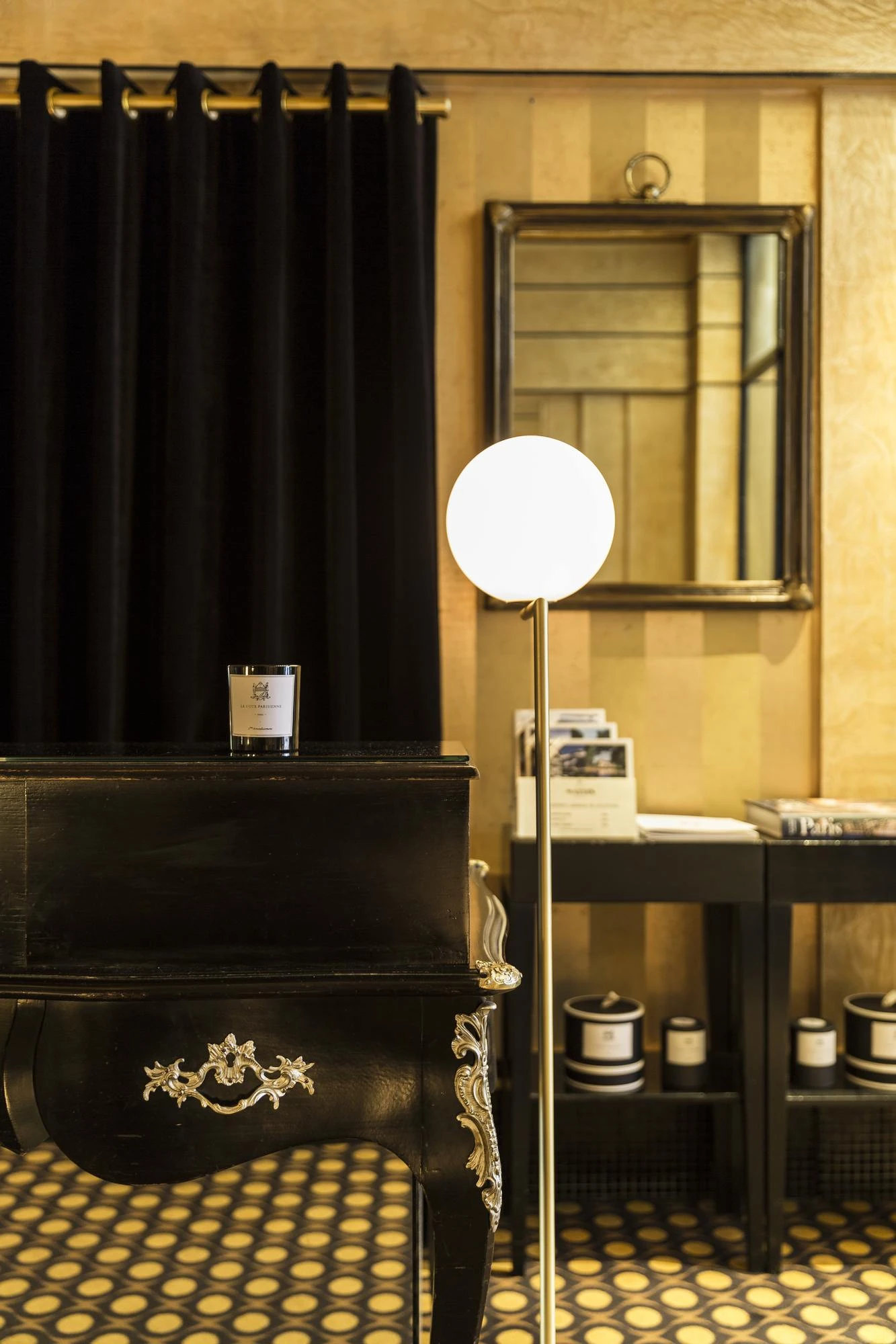 Intérieur élégant avec un meuble noir orné de détails dorés, une lampe sur pied à boule lumineuse blanche, un miroir encadré au mur et des tablettes noires contenant des bougies et des brochures, le tout sur un sol à motif circulaire jaune et noir.
