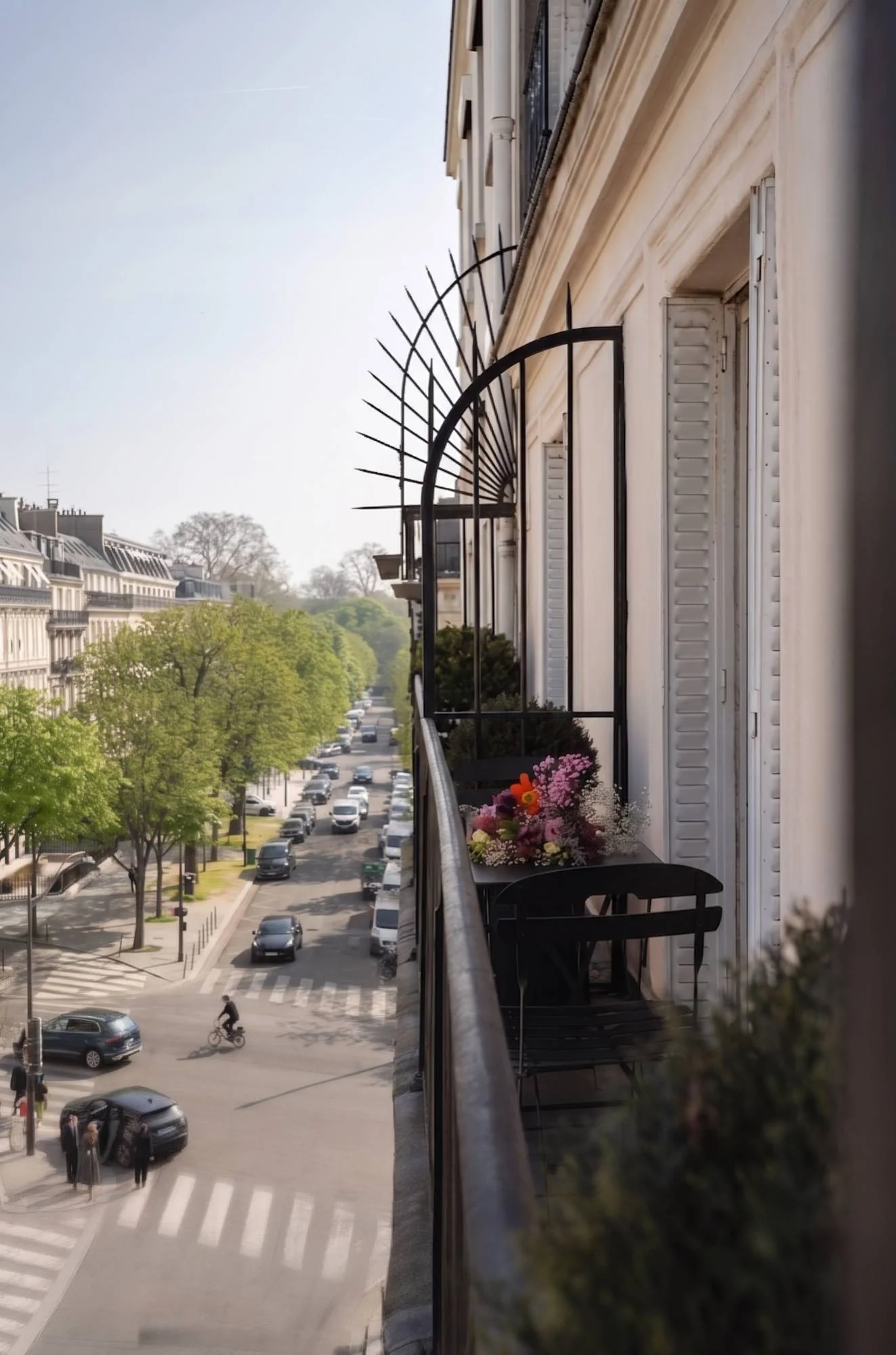 Balcon parisien avec une petite table noire, une chaise, et un bouquet coloré, donnant sur une rue bordée d'arbres et de voitures par une journée ensoleillée.