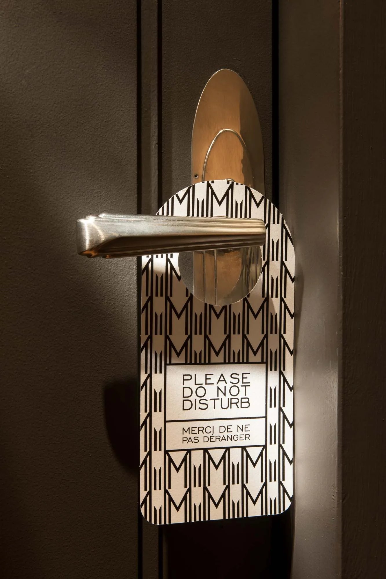 Une poignée de porte argentée avec un panneau suspendu indiquant "Please Do Not Disturb" et "Merci de ne pas déranger" en français, sur une porte sombre.
