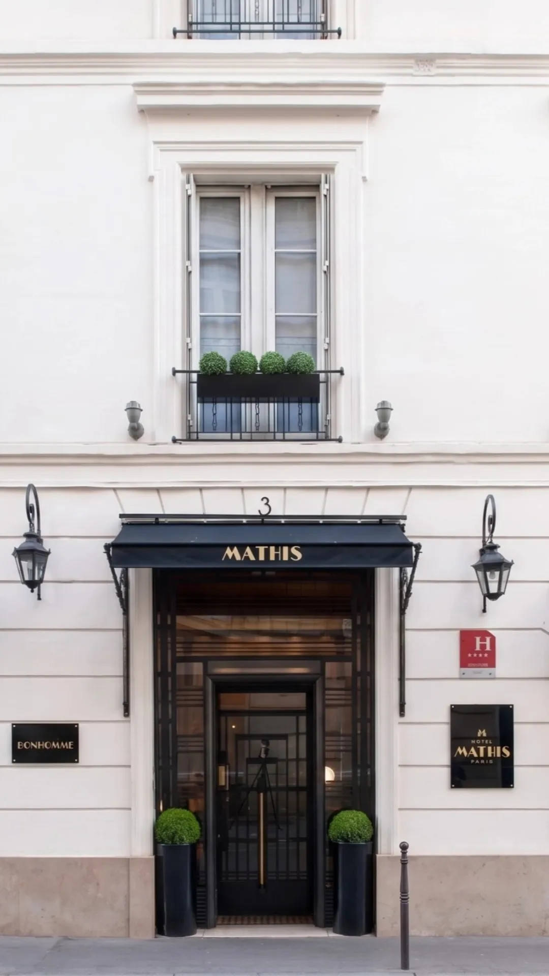 Façade élégante d'un hôtel parisien avec l'entrée sous une marquise noire portant l'inscription dorée « MATHIS », deux lanternes murales, des pots noirs avec des buissons verts de chaque côté de la porte, une fenêtre à volets blancs décorée avec quatre boules de verdure en jardinière, et des plaques indiquant « BONHOMME » et « HOTEL MATHIS PARIS » sur le mur blanc.