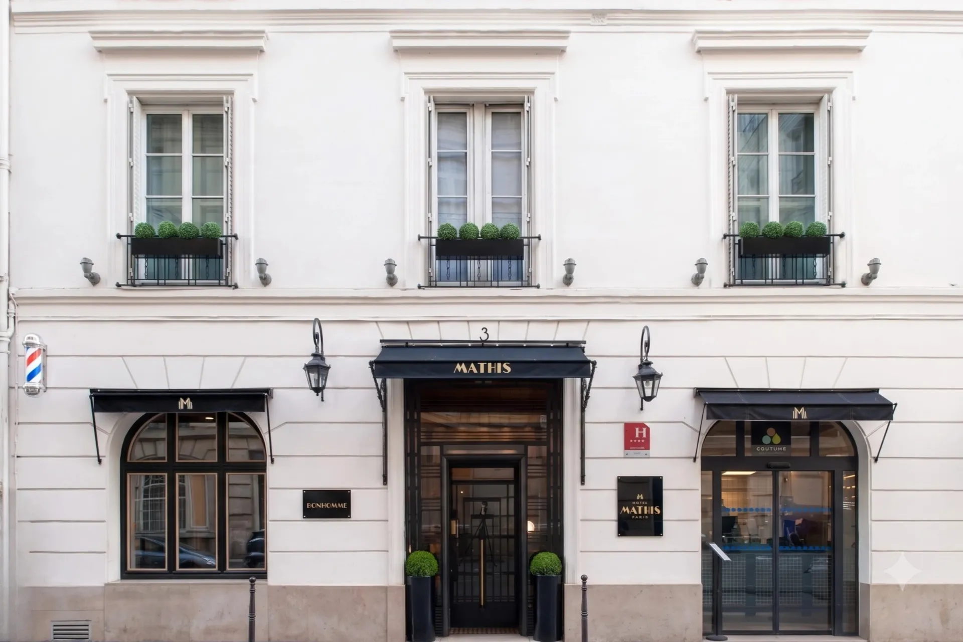 Façade blanche élégante d'un hôtel avec trois fenêtres à guillotine et un auvent noir au-dessus de l'entrée centrale marquée ‘Mathis’. Deux grandes jardinières noires avec des buissons verts encadrent la porte vitrée. Deux autres auvents noirs au-dessus des portes latérales portent le logo de l’hôtel. Un panneau rouge avec quatre étoiles indique la classification de l’hôtel à droite de l’entrée principale.