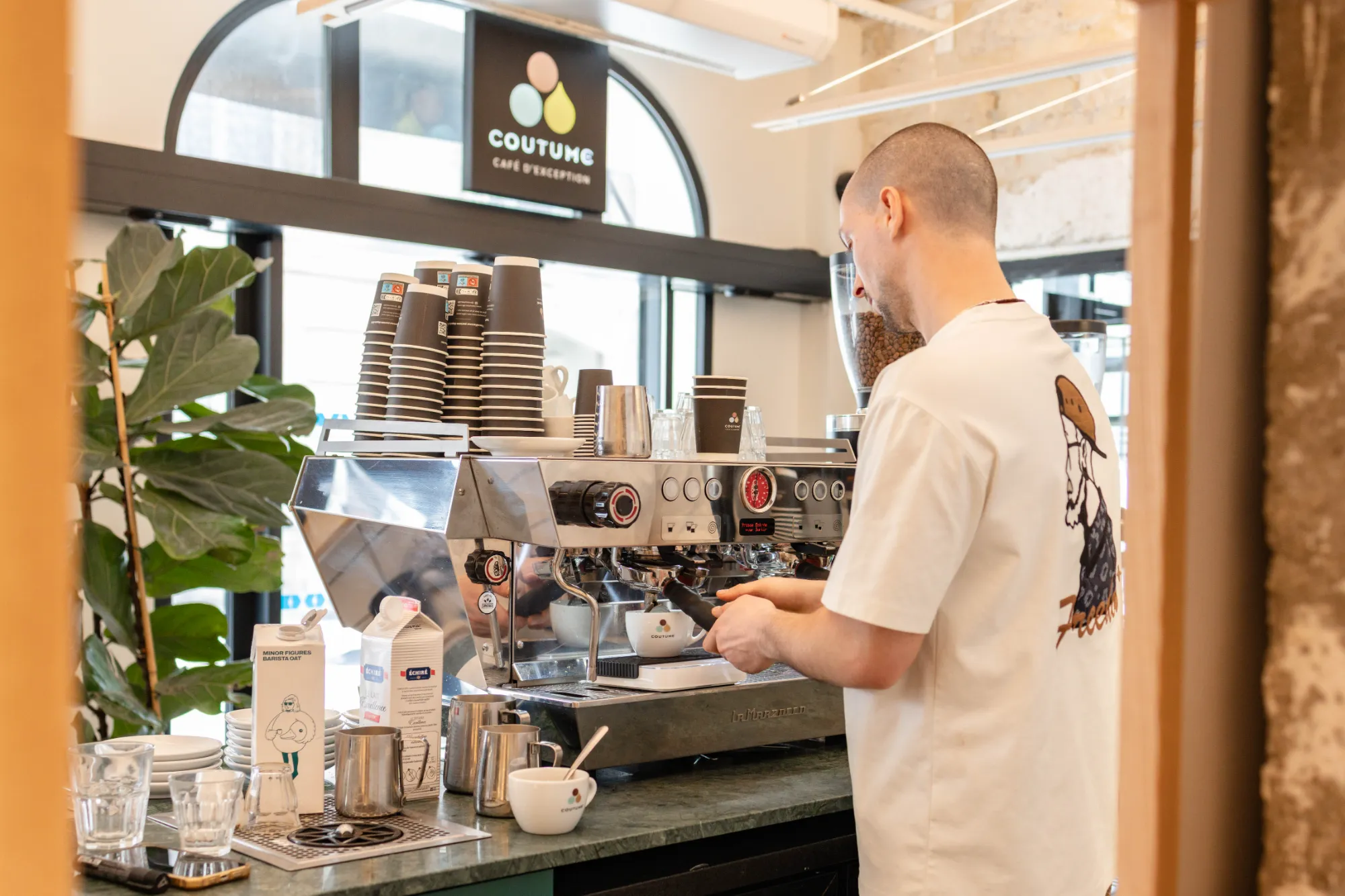 Barista préparant un café avec une machine à espresso La Marzocco dans un café lumineux avec une grande plante verte et une pile de gobelets en papier marqués Coutume.
