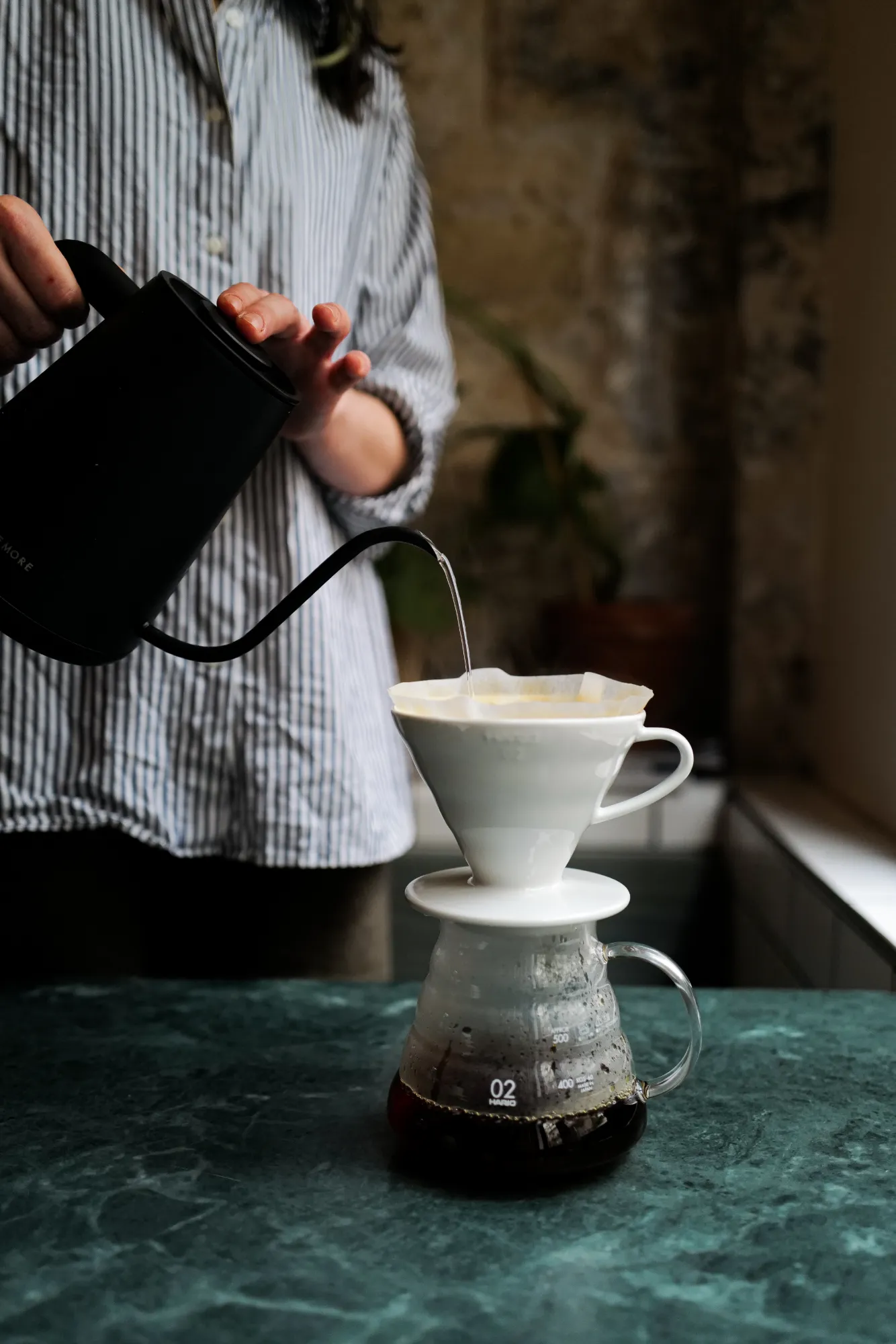 Une personne verse de l'eau chaude d'une bouilloire noire à bec long dans un filtre en papier blanc placé sur un support pour café filtre, avec une cafetière en verre recueillant le café infusé sur une table en marbre vert.