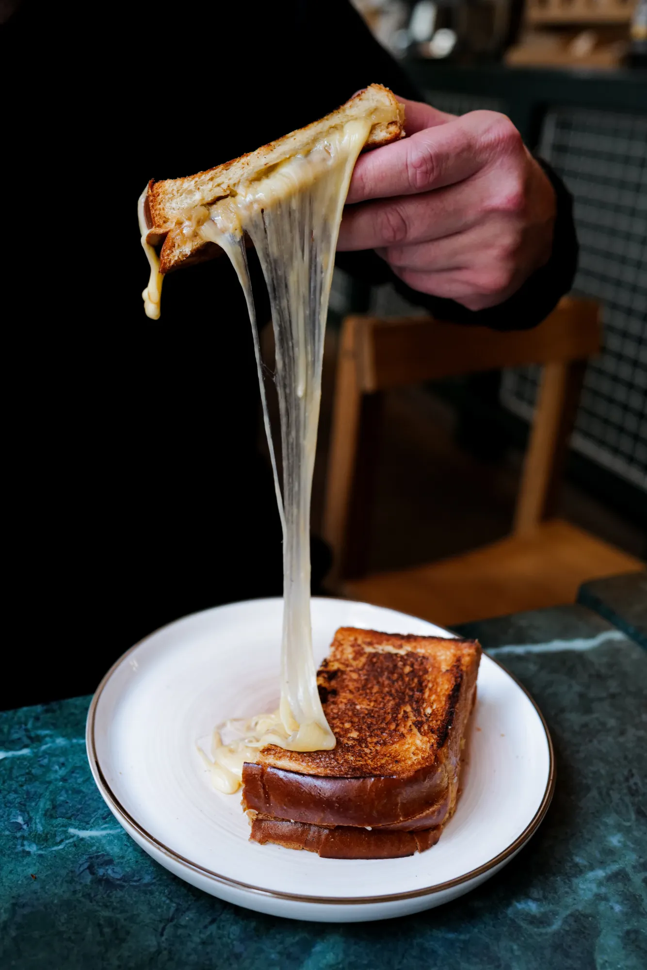 Une main tient un sandwich grillé au fromage fondant, tirant le fromage qui s’étire en longs filaments au-dessus d’une assiette blanche sur une table en marbre vert.