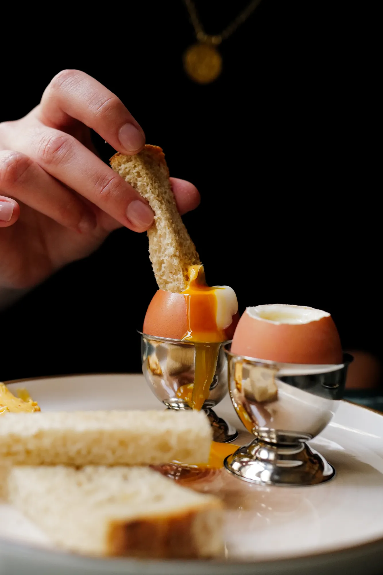 Une main trempe un morceau de pain dans un œuf à la coque dont le jaune coule légèrement hors de la coquille posée dans un coquetier en métal, un autre œuf à la coque est placé à côté, sur une assiette avec des tranches de pain.
