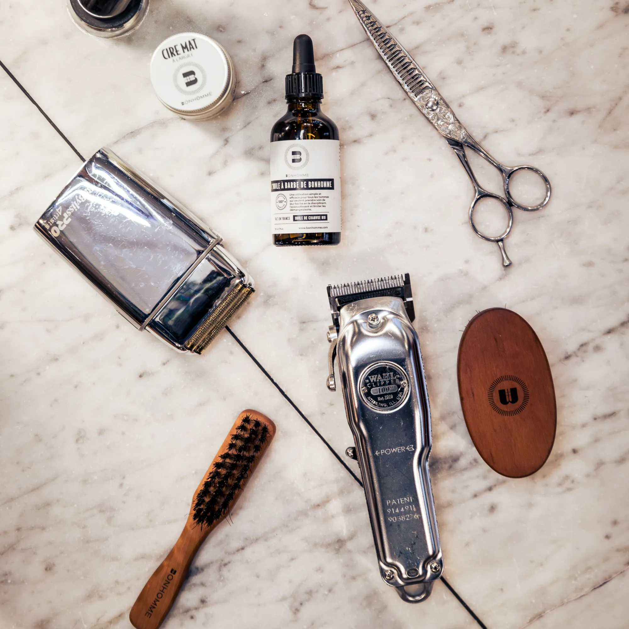 Ensemble d'outils de barbier sur un fond en marbre, comprenant tondeuse électrique, ciseaux, brosse à poils sombres, huile à barbe dans une bouteille avec compte-gouttes, cire mate en pot, rasoir électrique et une brosse ovale en bois.