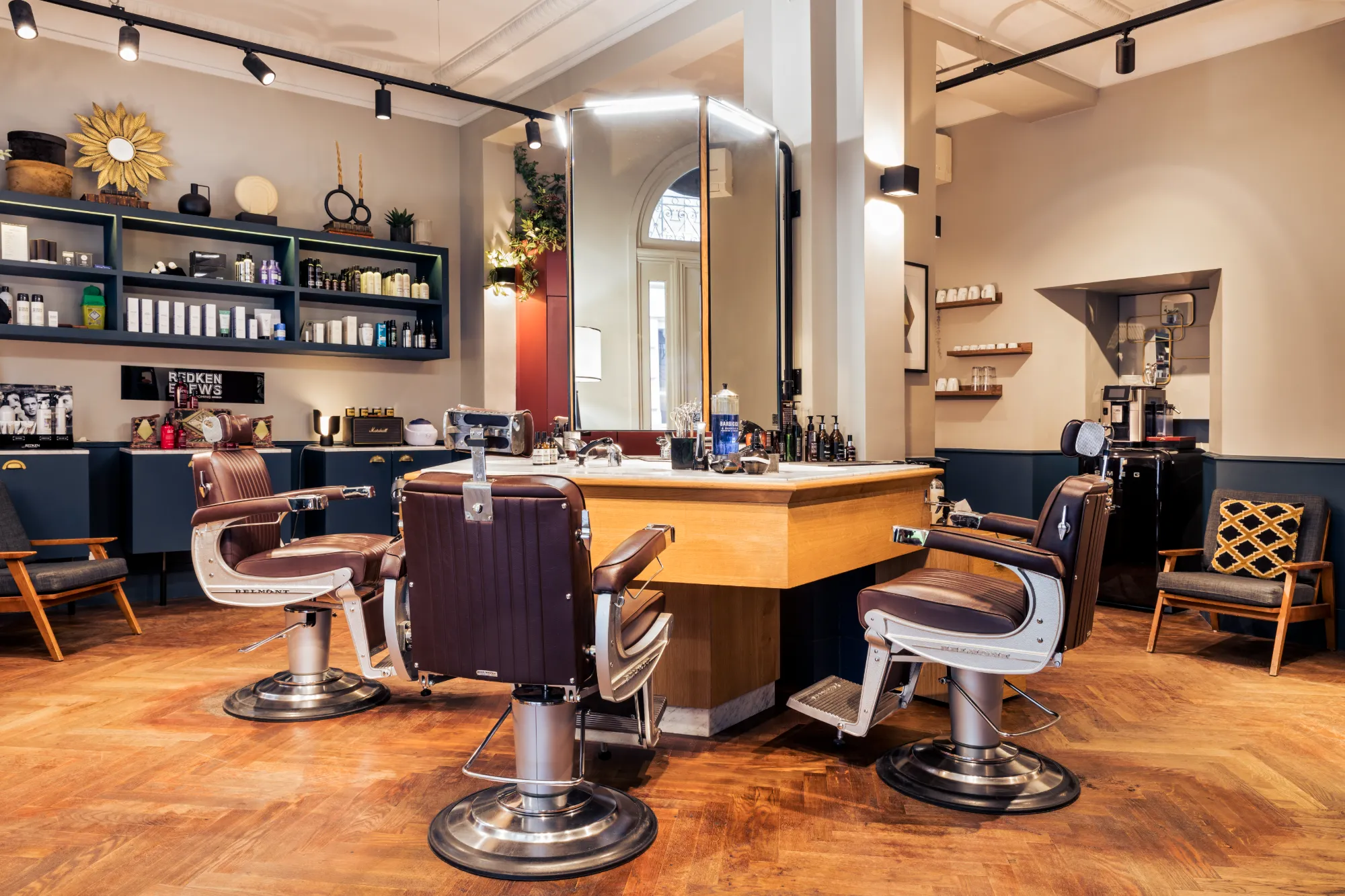 Intérieur d'un salon de coiffure moderne avec trois fauteuils de barbier en cuir marron autour d'un grand miroir central sur un comptoir en bois, étagères avec produits de soins, et un coin café avec fauteuils en bois.
