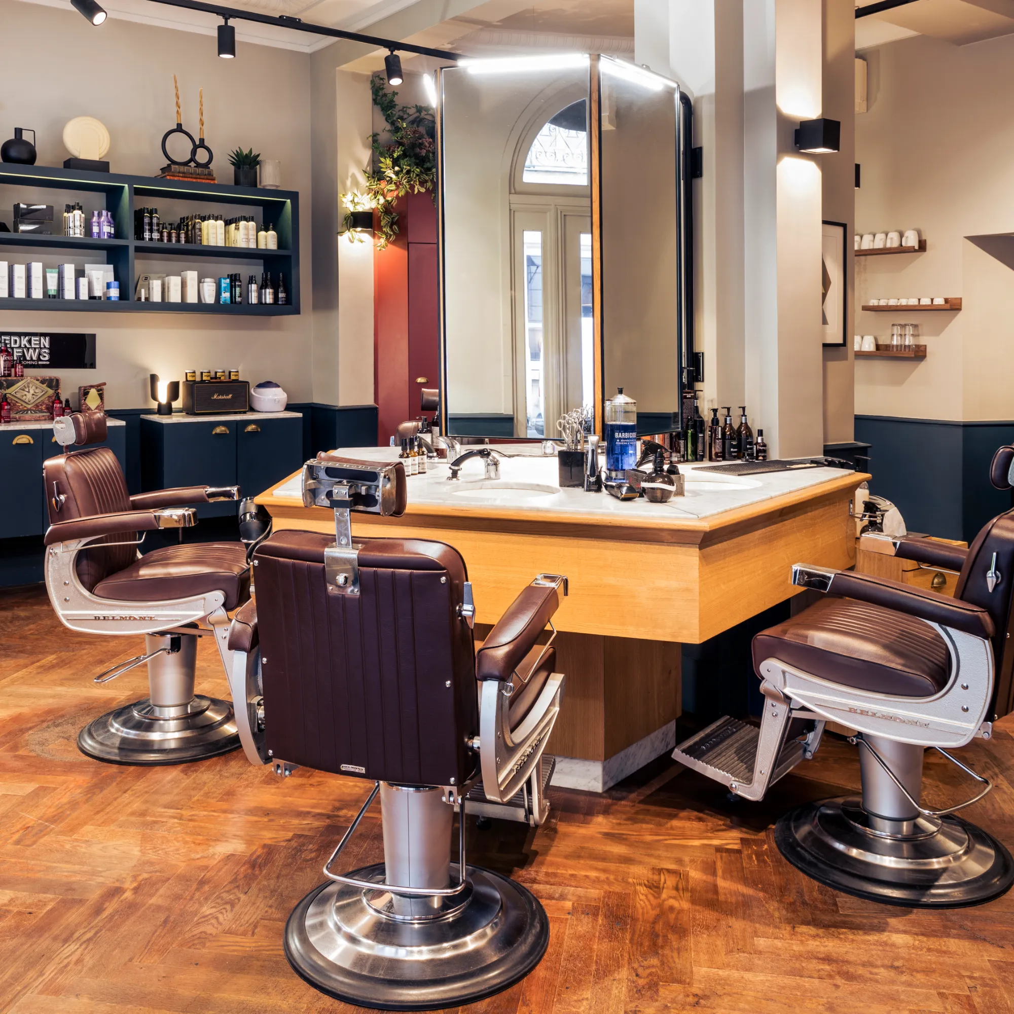 Salon de barbier élégant avec trois fauteuils en cuir marron autour d'une grande coiffeuse en bois avec miroir sur mesure, contenant des produits de soin et accessoires de coiffure, le tout sur un parquet en bois foncé.