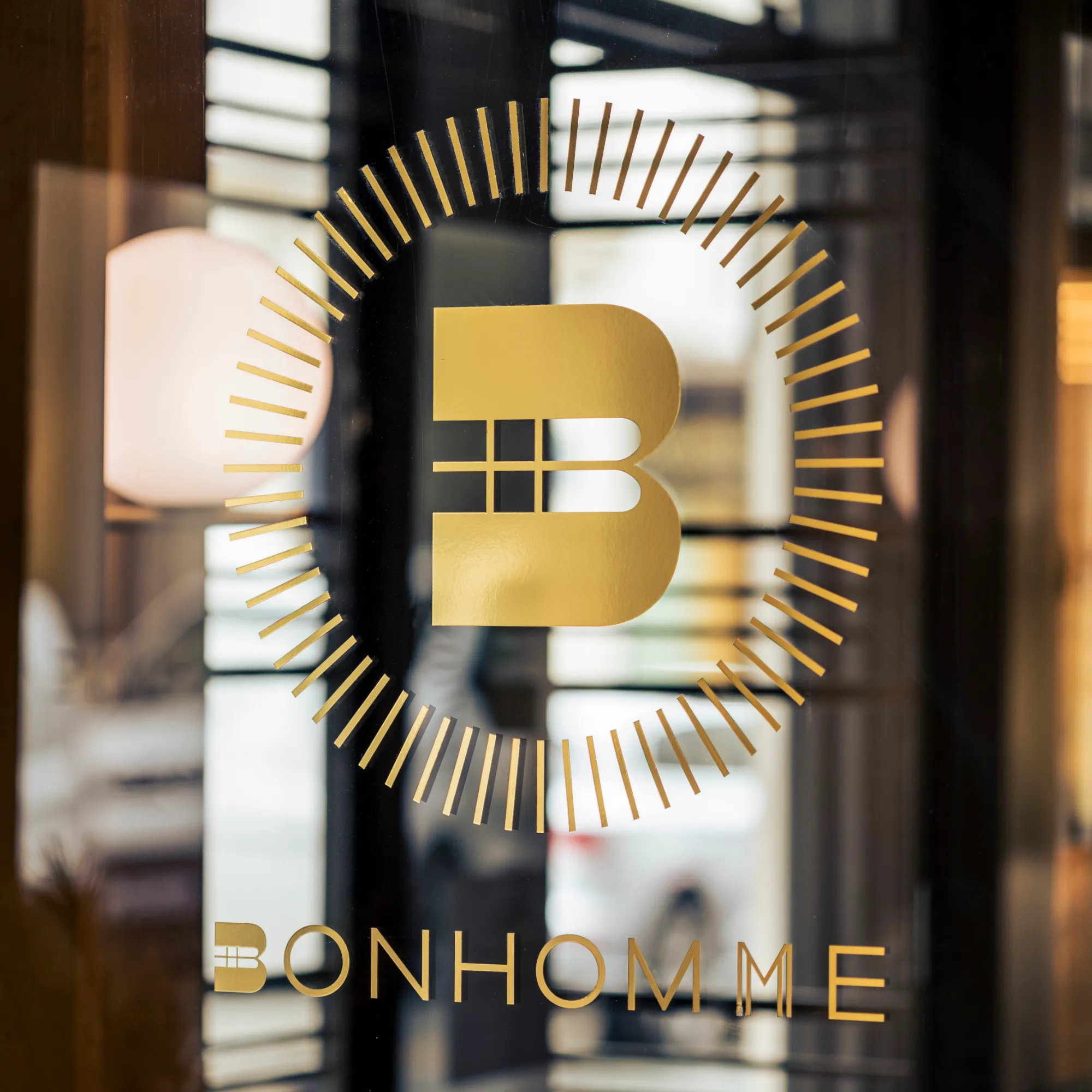 Logo doré stylisé avec un grand B entouré de traits dorés formant un cercle et le mot BONHOMME en dessous sur une porte en verre reflétant l'intérieur d'un bâtiment.