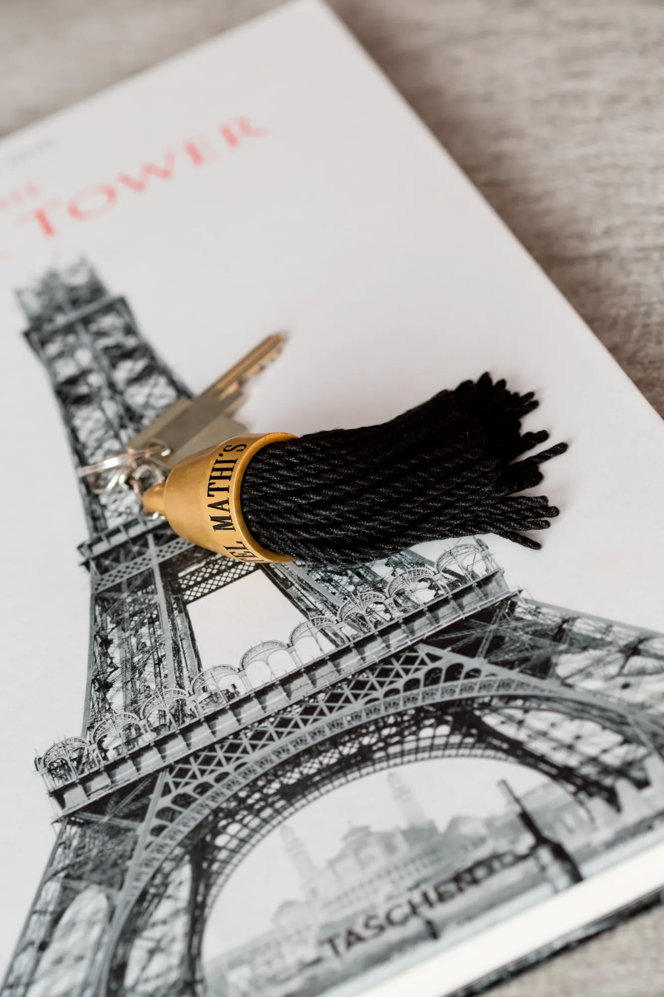 Clé avec un porte-clés à pompon noir et anneau doré marqué "El Mathi's" posée sur un livre illustré de la Tour Eiffel.