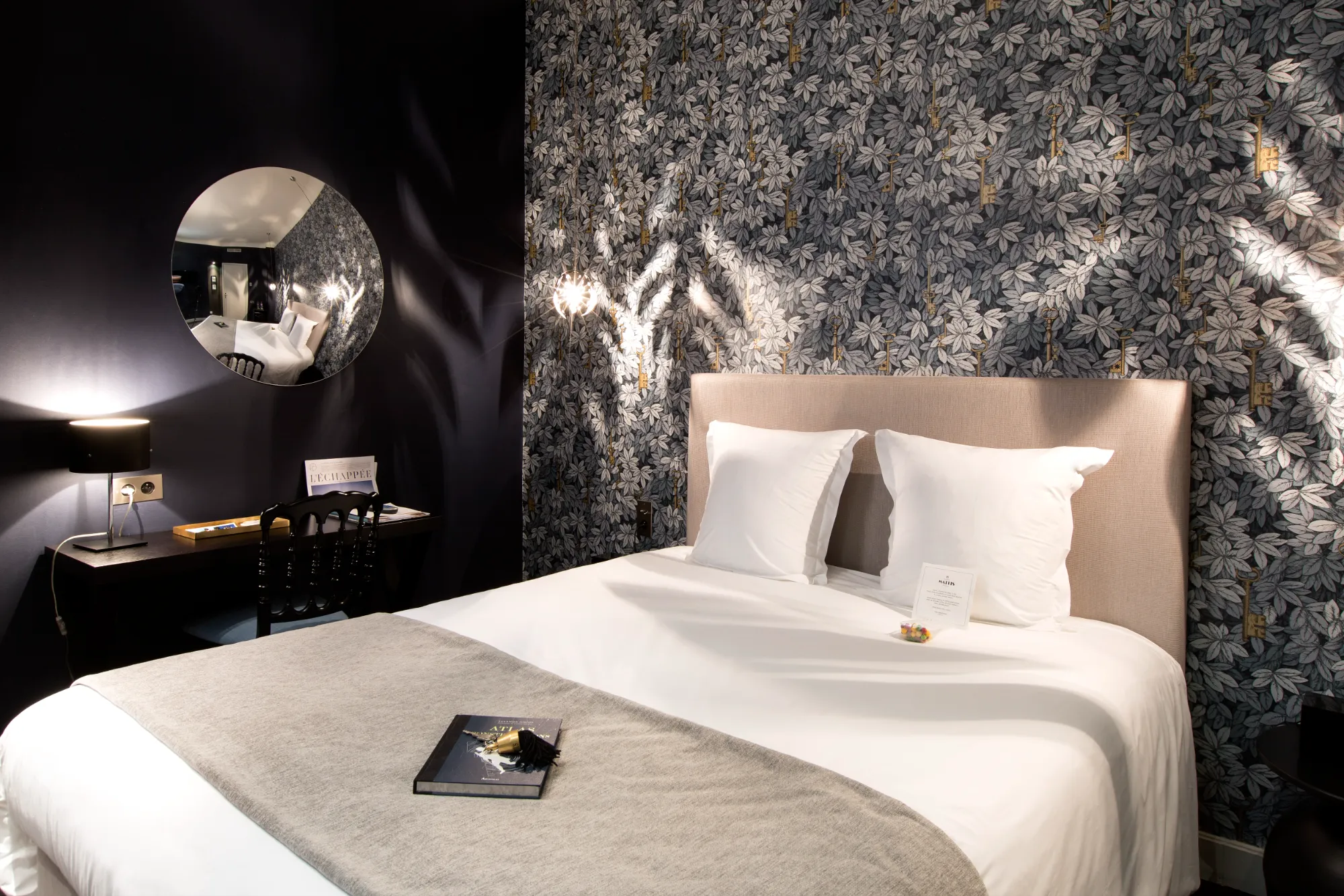 Chambre d'hôtel avec un grand lit double à tête beige, deux oreillers blancs et un plaid gris clair. Un livre avec un trousseau de clés est posé sur le lit. Le mur derrière le lit est décoré d'un papier peint à motifs de feuilles grises et de clés dorées. À gauche, un bureau noir avec une lampe allumée, une chaise noire, et un miroir rond reflétant le lit et le papier peint. Lumière douce créant des ombres délicates sur les murs.
