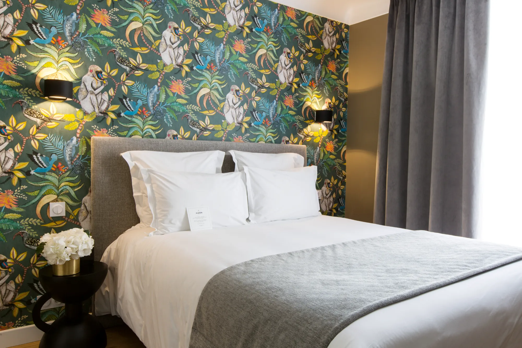 Chambre d'hôtel avec un grand lit double aux draps blancs et un plaid gris, tête de lit grise, murs ornés de papier peint coloré avec motifs de singes et oiseaux, deux appliques lumineuses noires et une petite table noire avec un vase de fleurs blanches, rideaux gris occultants à droite.