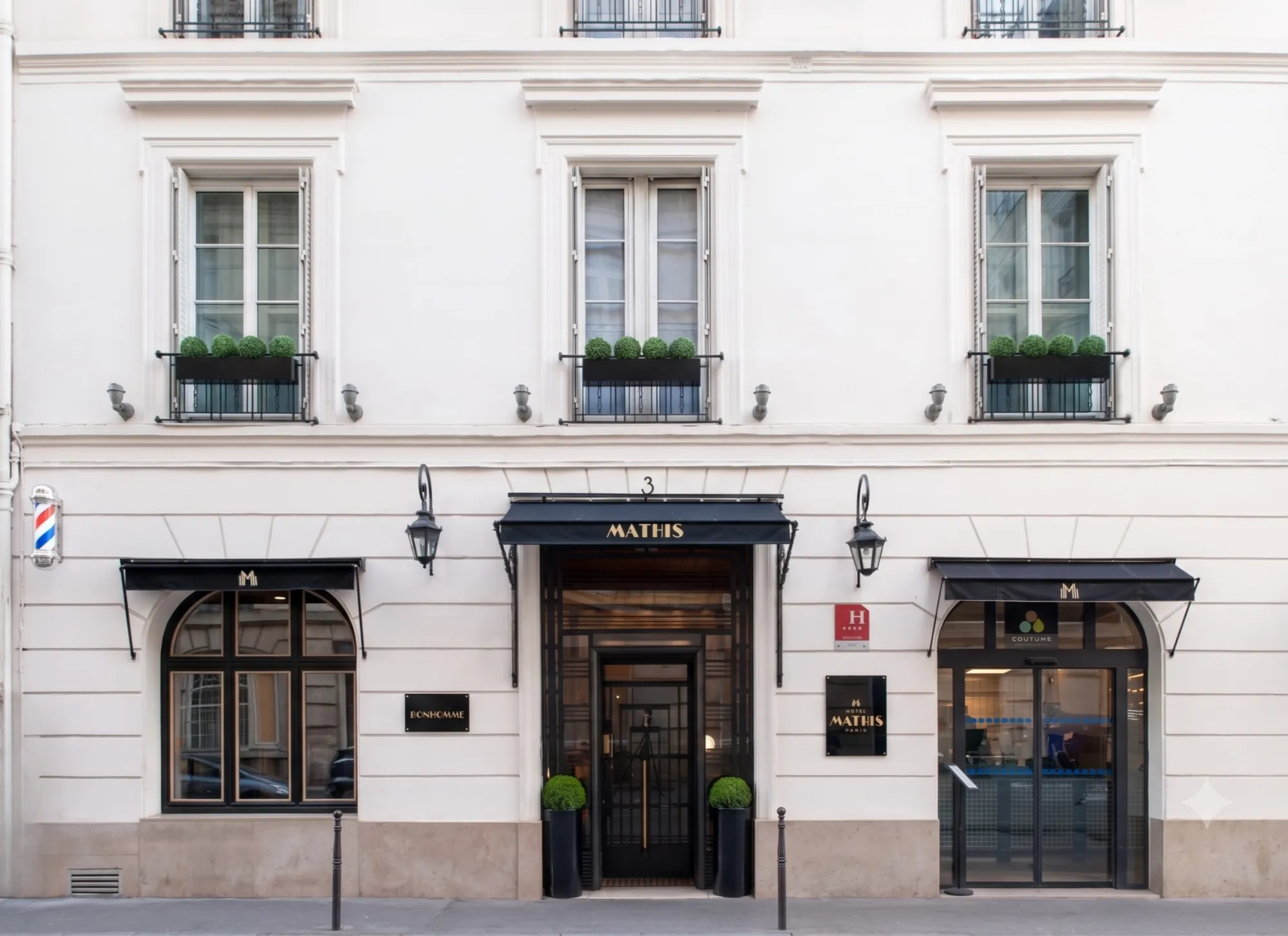 Façade blanche élégante d'un bâtiment parisien avec trois fenêtres, chacune décorée d'une jardinière de buissons ronds. Au centre, une entrée noire avec un auvent portant l'inscription "MATHIS" en lettres dorées. À gauche, une fenêtre sous un autre petit auvent noir à côté d'un panneau qui dit "BONHOMME". À droite, une porte vitrée sous un auvent noir conduit à "COUTUME", un espace intérieur visible. Deux grands pots avec des buissons verts encadrent la porte centrale.