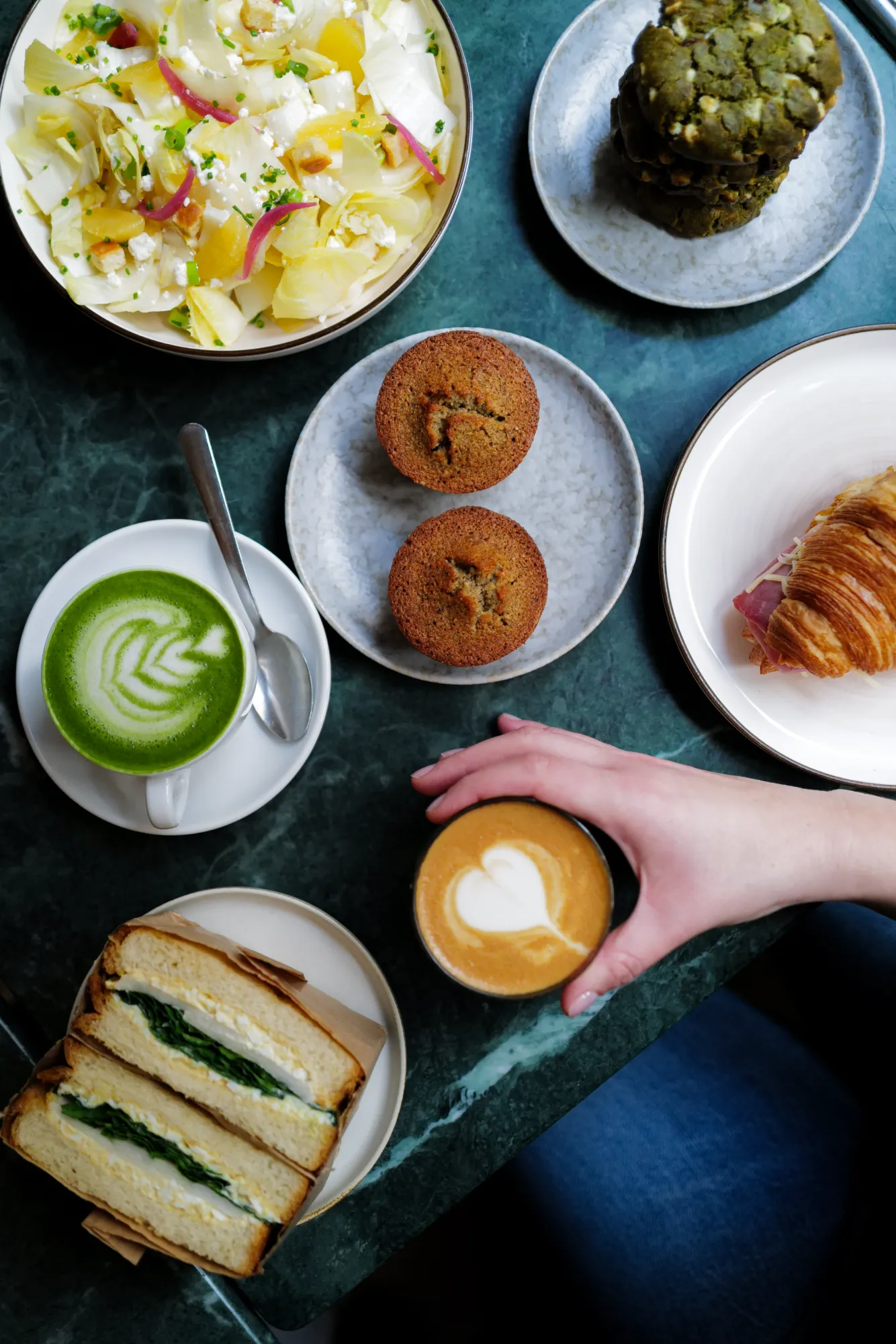 Vue du dessus d'un repas sur une table en marbre vert incluant une salade d'endives, des muffins, des cookies verts, un croissant avec jambon, un sandwich aux œufs et épinards, une tasse de café latte avec art latte en forme de cœur tenue par une main, et une tasse de matcha latte avec une cuillère sur la soucoupe.