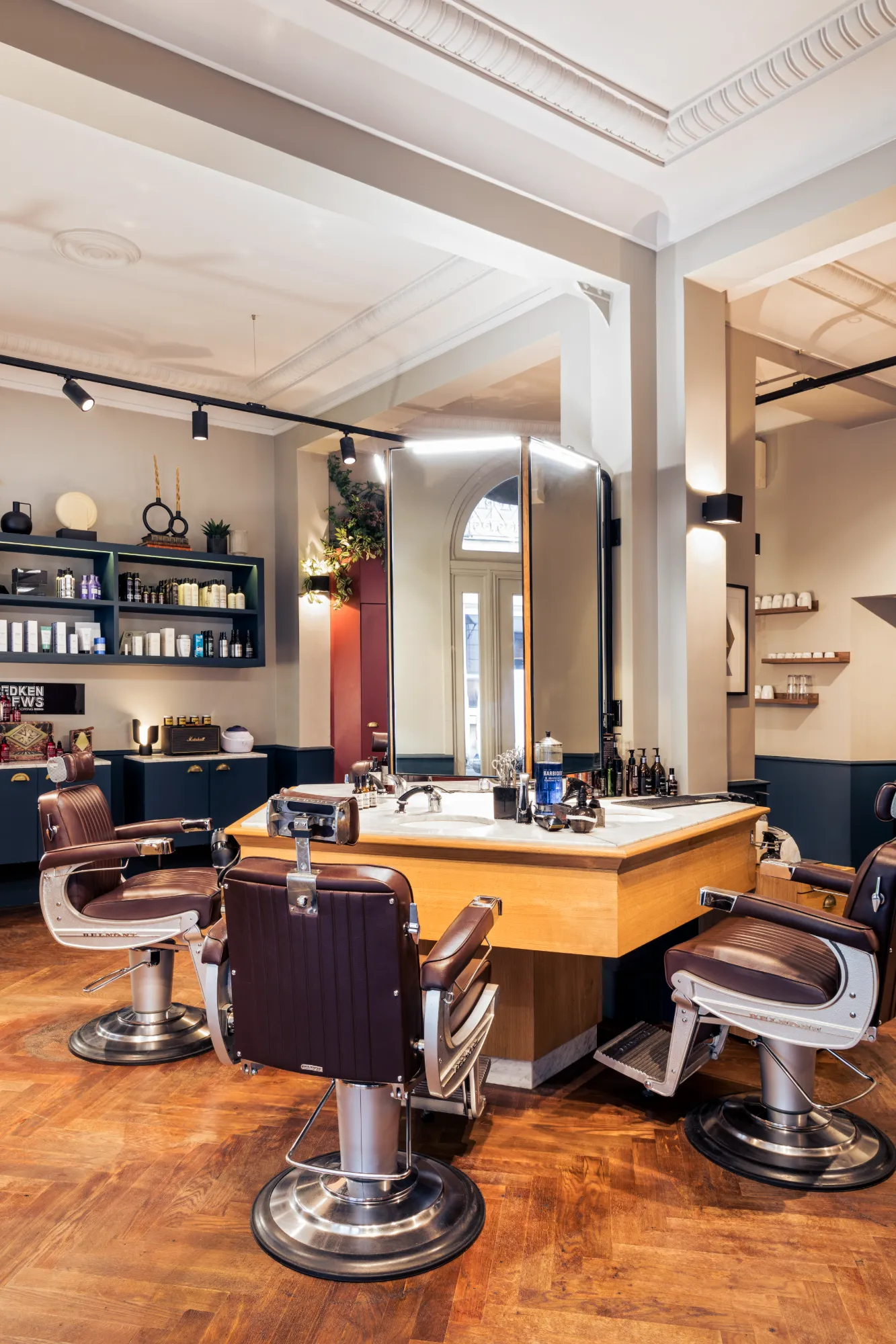 Intérieur d'un salon de barbier moderne avec trois chaises de barbier en cuir marron autour d'un comptoir en coin équipé d'un grand miroir, des produits coiffants et des outils de barbier soigneusement disposés, parquet en bois et murs décorés de plusieurs étagères avec des produits de soins.