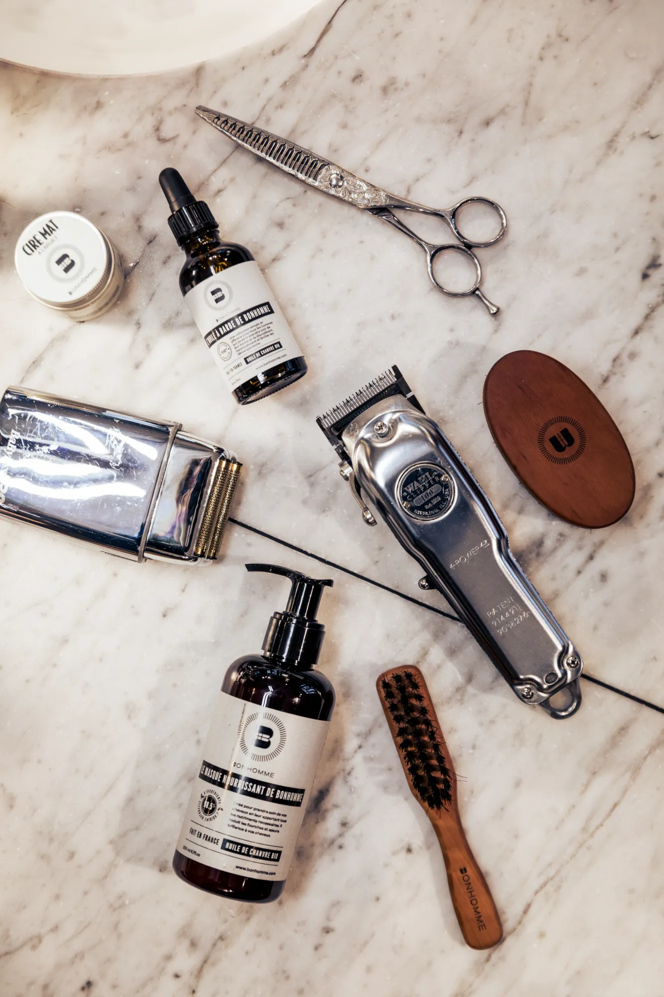 Ensemble d'outils de barbier sur surface en marbre, incluant une paire de ciseaux de coiffure, une tondeuse électrique chromée avec câble, un peigne en bois avec poils noirs, un flacon pompe de soin pour barbe, un flacon compte-gouttes d'huile pour barbe, un petit pot de crème et une brosse ovale en bois.