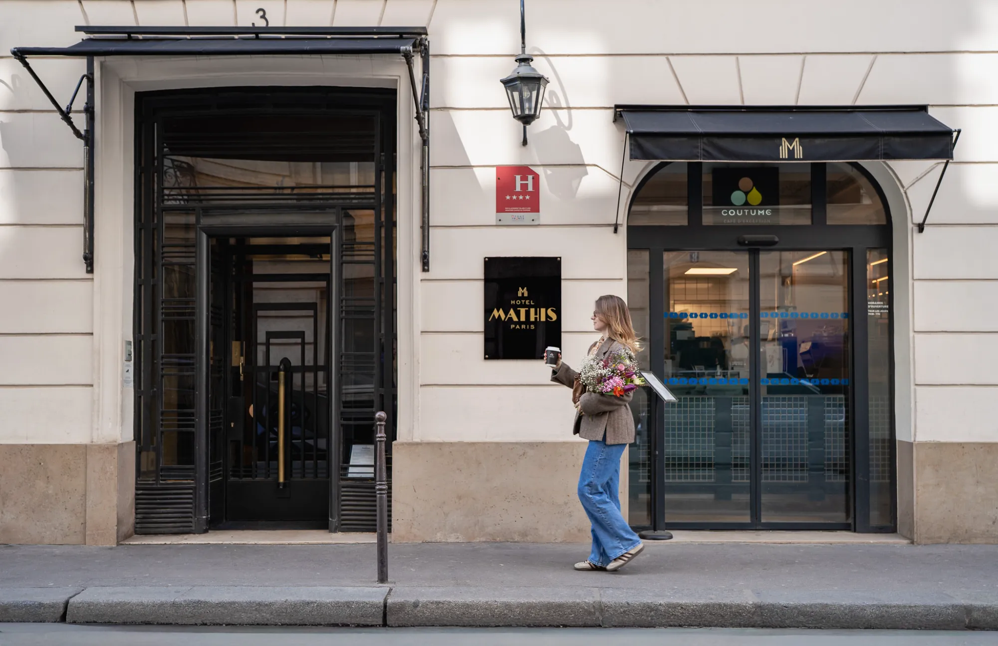 Une femme marche devant l'entrée de l'Hôtel Mathis à Paris, tenant un bouquet de fleurs et une boisson à emporter, avec une autre porte d'entrée à droite montrant un café nommé Coutume.