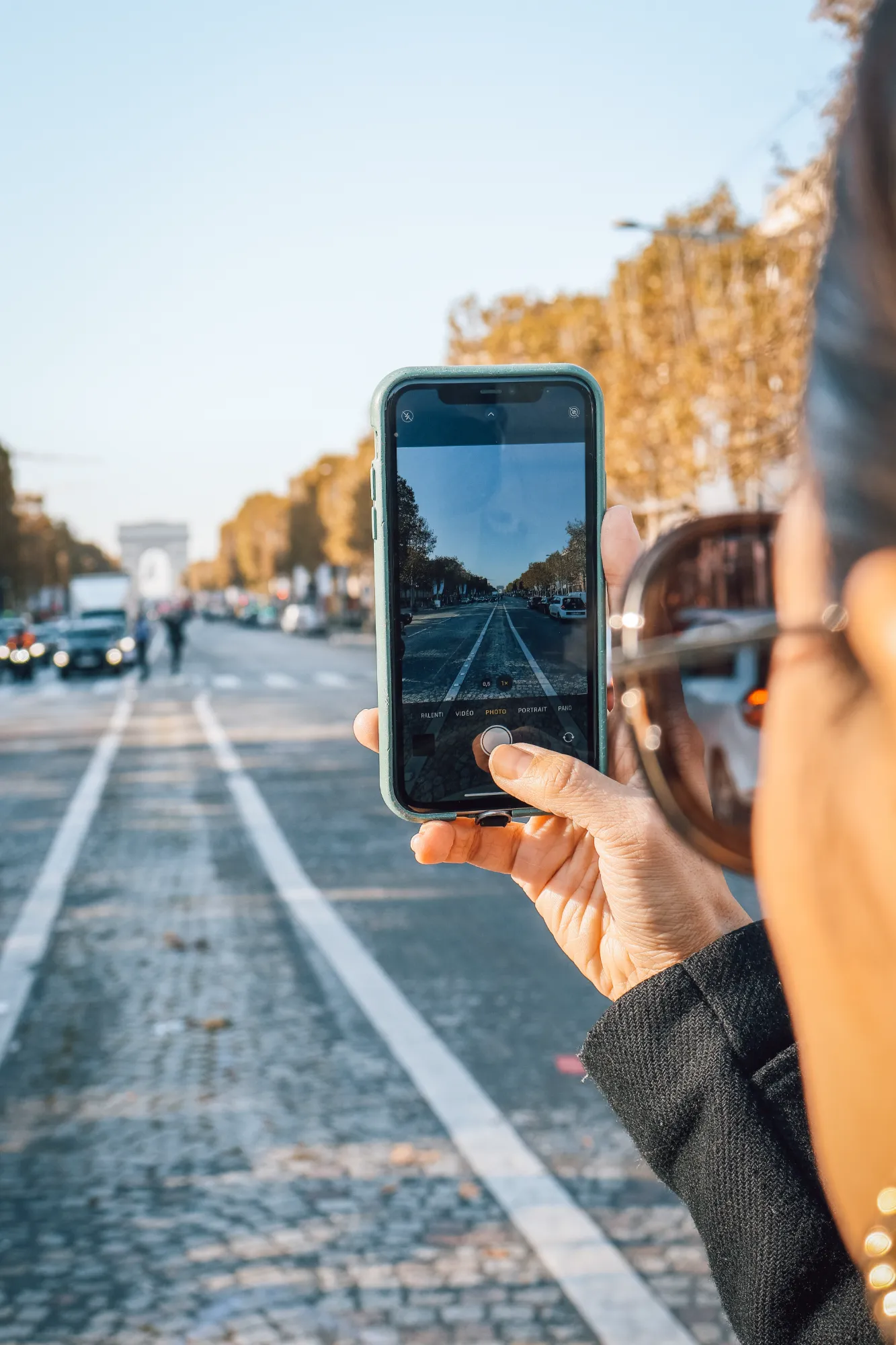 Person tenant un smartphone montrant en photo l'avenue des Champs-Élysées avec l'Arc de Triomphe en arrière-plan flou, sous un ciel dégagé.