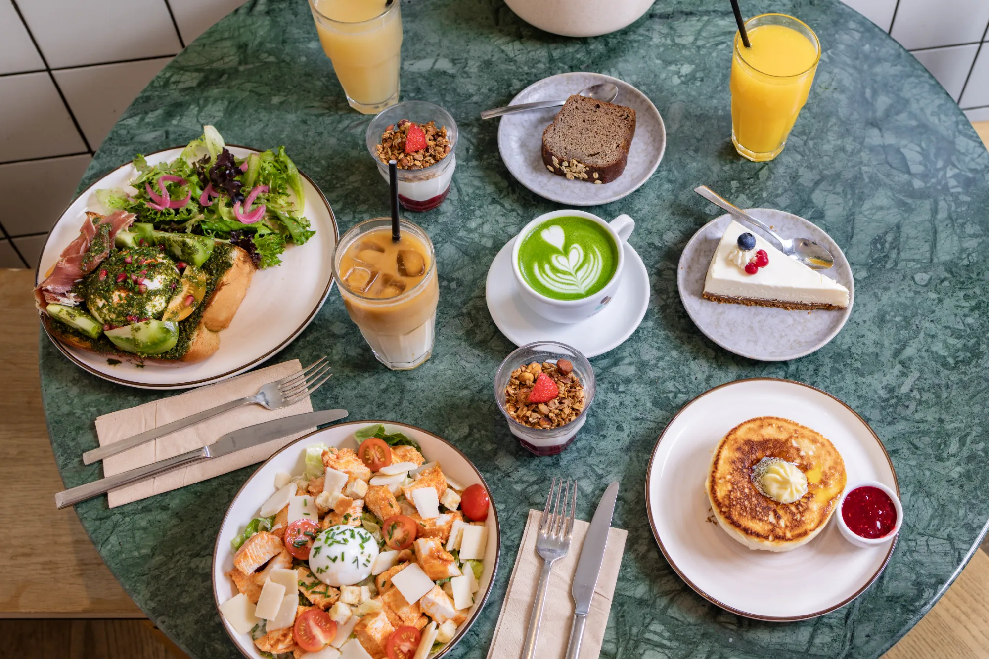 Vue de dessus d'une table ronde verte avec un brunch composé de deux salades avec œufs pochés et tomates, deux verres de jus d'orange, deux yaourts granola avec fraise, un café matcha avec décoration en mousse, une tranche de pain brun, une part de gâteau blanc aux baies, un verre de café glacé à la paille, et une assiette avec deux pancakes, une noix de beurre et un petit bol de confiture rouge.