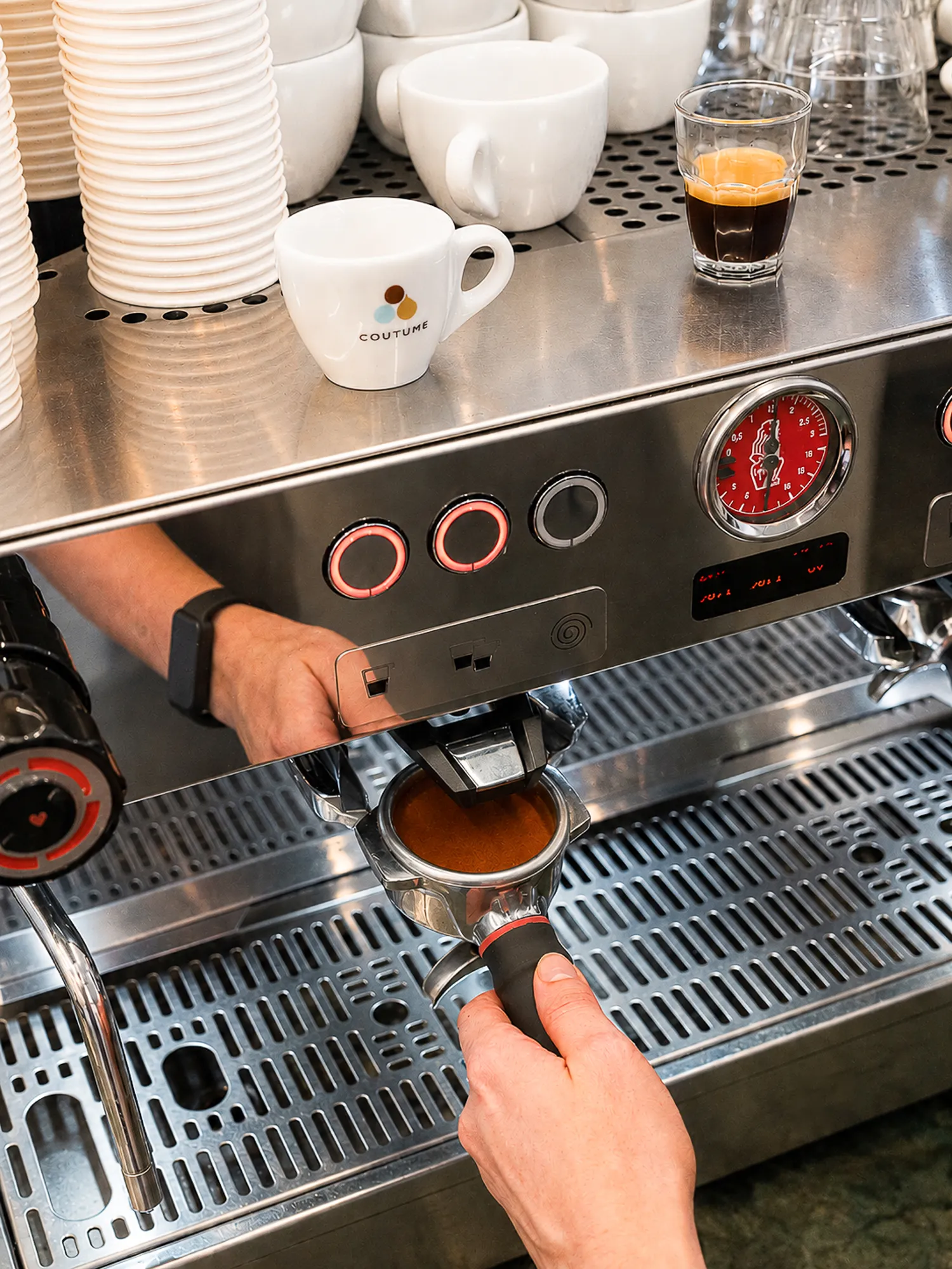 Une machine à expresso en acier inoxydable avec une main tenant un porte-filtre pendant l'extraction d'un café espresso. Sur le dessus de la machine sont posées plusieurs tasses en porcelaine blanche, dont une avec le logo Coutume, et un verre contenant un expresso avec une crème onctueuse.