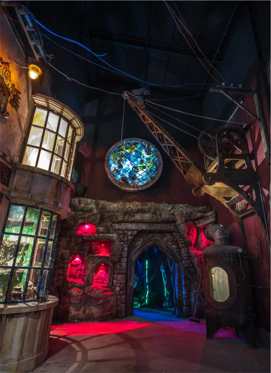 Fantasy-themed interior with stone archway, red and blue lighting, stained glass window, and vintage industrial machinery in a dimly lit immersive exhibit.