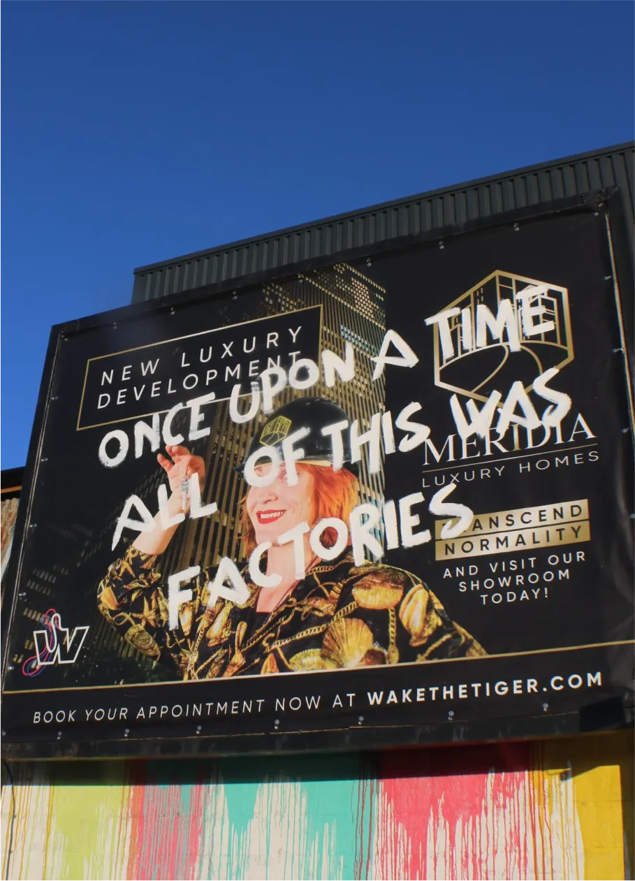 Billboard for luxury homes with graffiti text reading “Once upon a time all of this was factories” over an image of a smiling woman in a gold-patterned outfit.