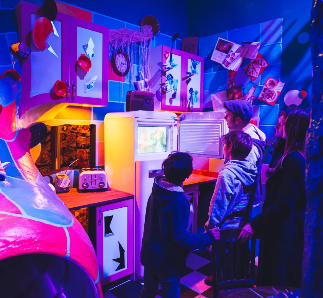 Family exploring a vibrant, surreal kitchen installation with neon lighting, open fridge, and whimsical decorations.