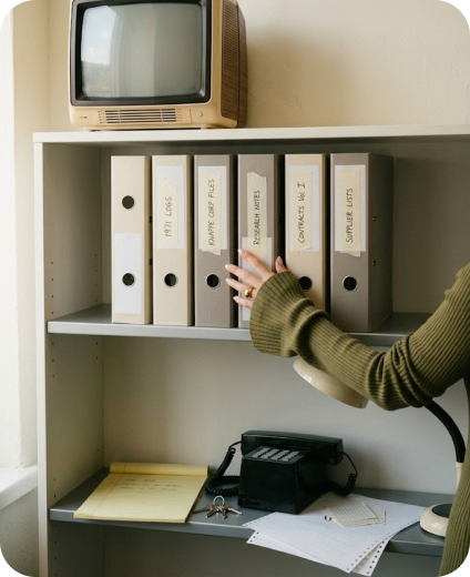 Hand reaching for labeled archive binders on a shelf beside an old TV and rotary phone