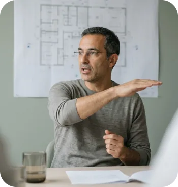 Man in glasses gesturing while explaining in front of floor plans pinned to a wall
