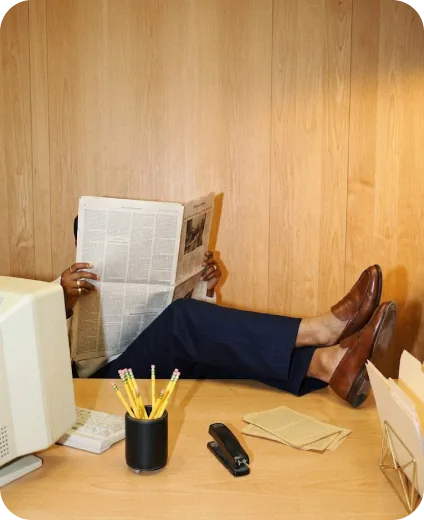Person hidden behind a newspaper with feet up on a desk beside a vintage computer