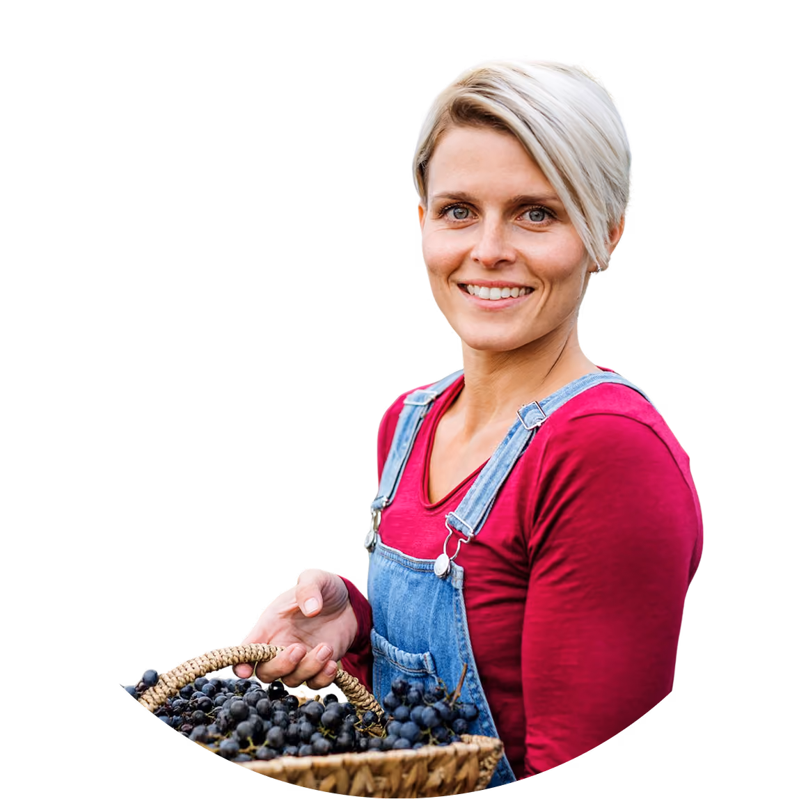 Smiling woman with short blonde hair holding a basket of dark grapes, wearing denim overalls and red shirt.