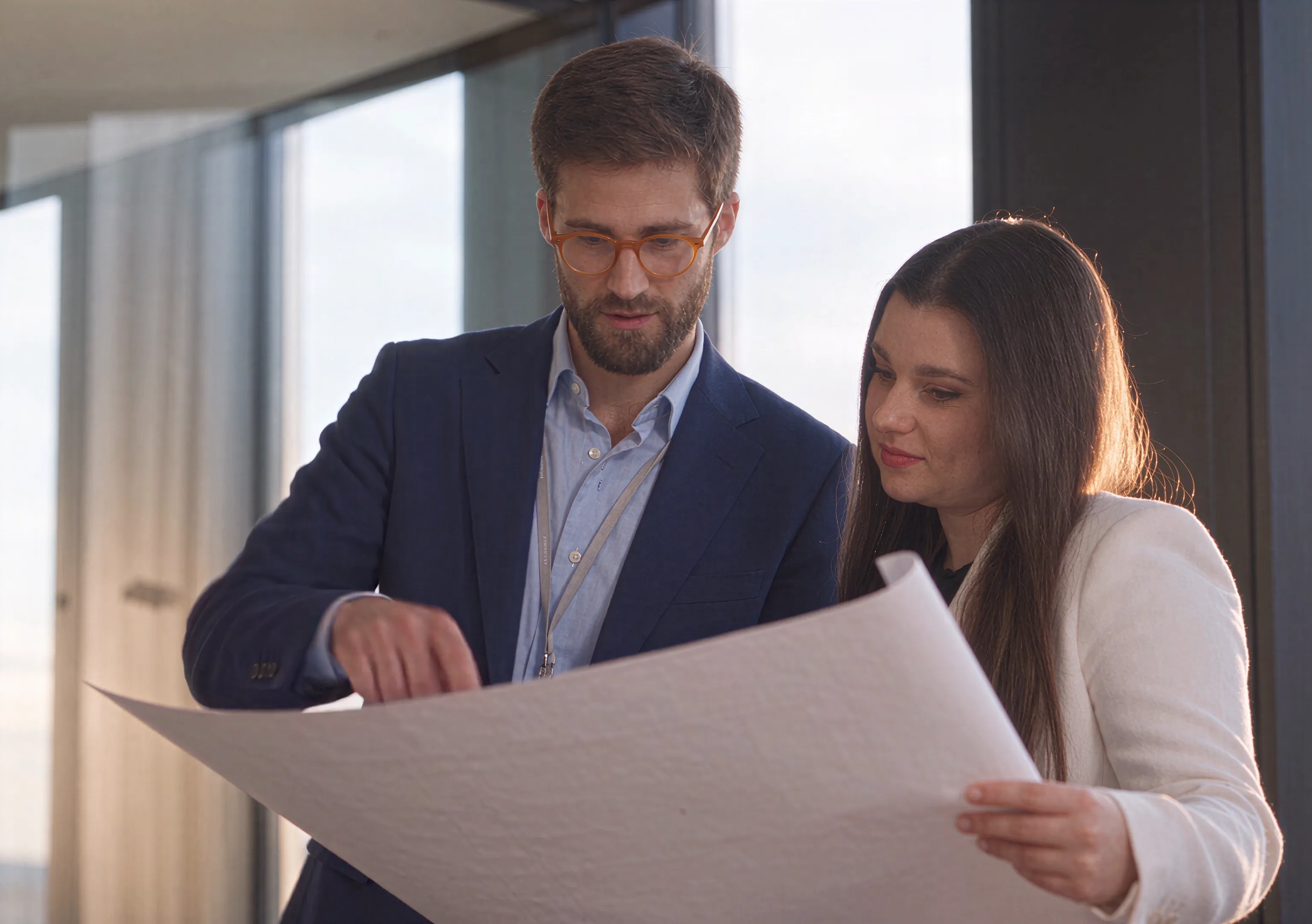 Two colleagues reviewing a large printed drawing in a glass-walled office