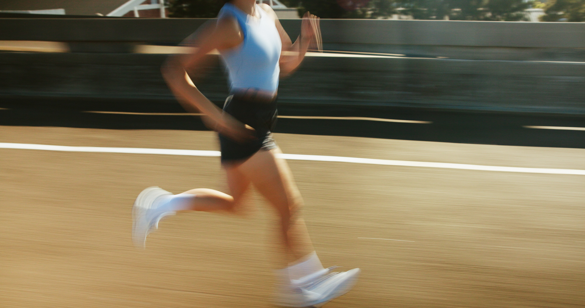 Blurred image of a person running on a road wearing a light blue sleeveless top, black shorts, and white sneakers, captured in motion against a concrete barrier background.