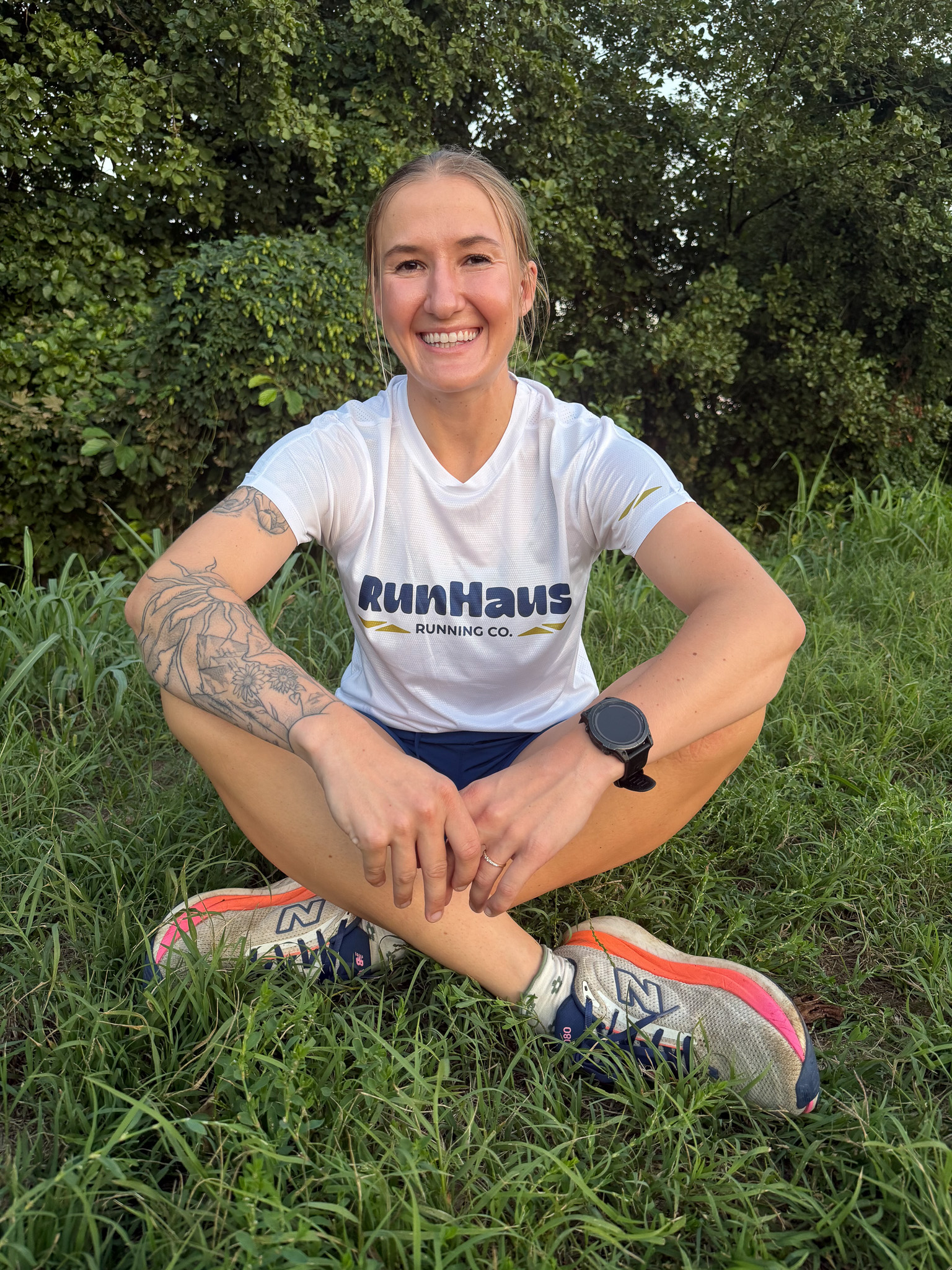 Smiling person sitting cross-legged on grass wearing a white RunHaus Running Co. shirt, blue shorts, New Balance running shoes, a black watch, and showing a tattoo on their left arm with greenery in the background.