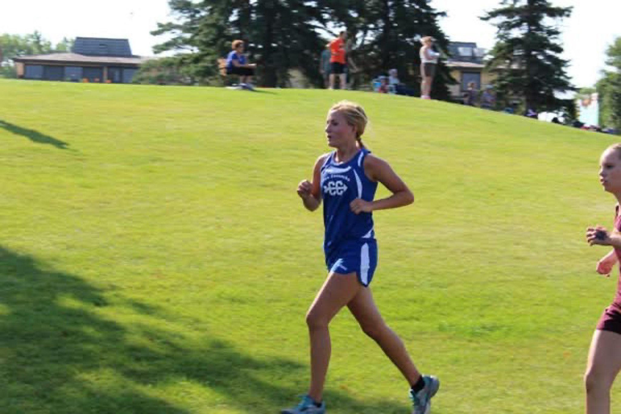 A young female runner in a blue cross country singlet and shorts running on a grassy slope with trees and several people in the background.