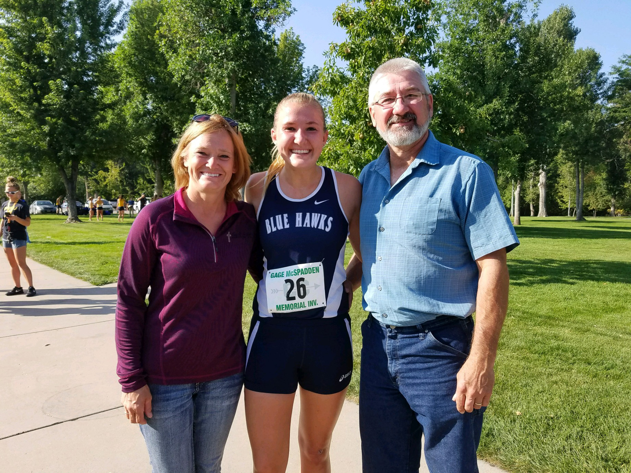 Three people smiling and posing outdoors on a sunny day in a park-like setting, with green trees and grass. The person in the middle is a young female athlete wearing a Blue Hawks athletic uniform and a race number 26, flanked by a woman in a maroon zip-up jacket and jeans on the left, and a man in a light blue short-sleeve shirt and jeans on the right.