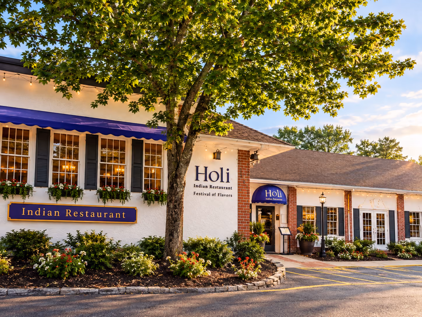 Exterior of Holi Indian Restaurant with white walls, flower beds, and a large tree in front.
