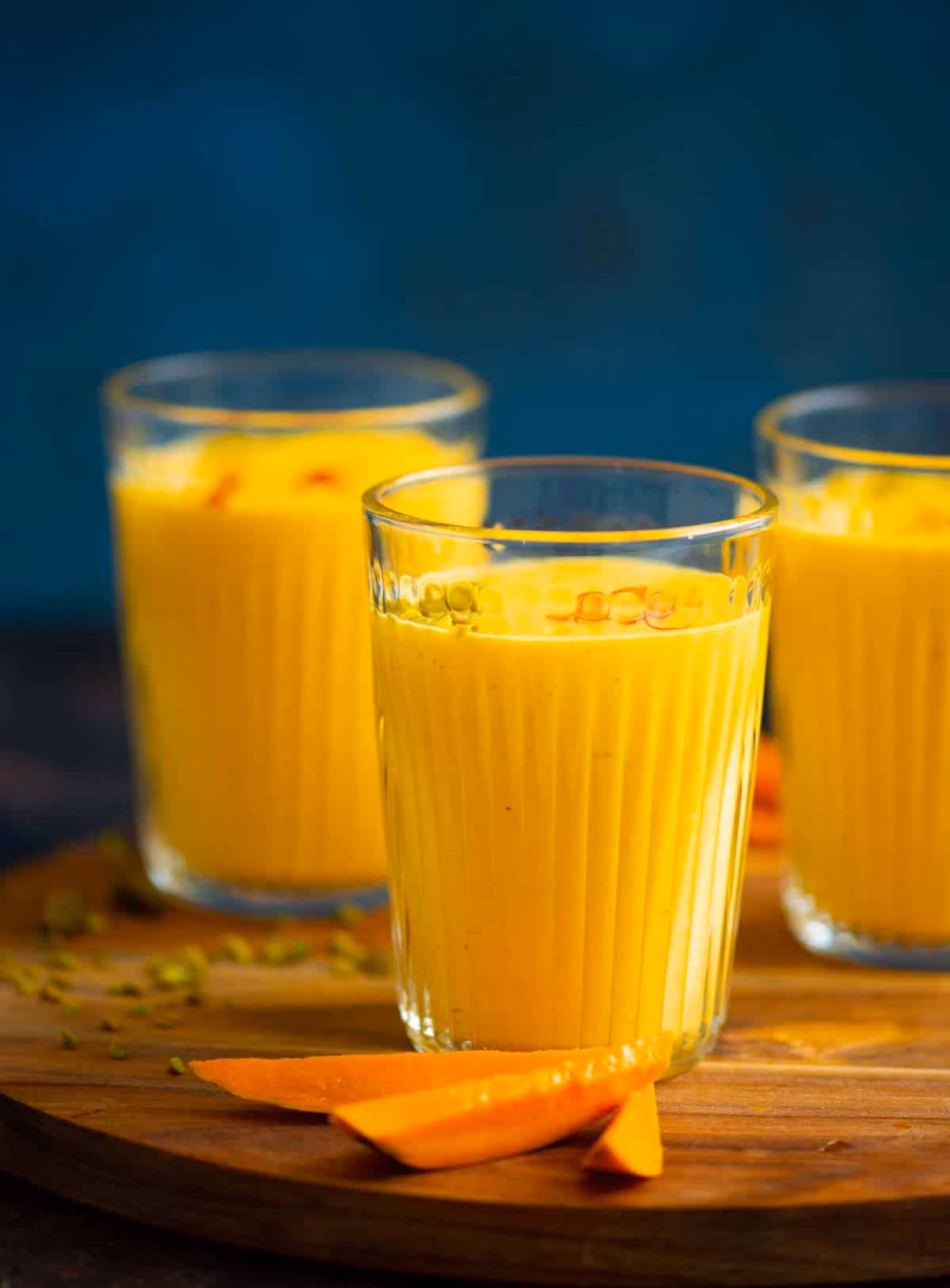 Three glasses of yellow mango lassi on a wooden board with mango slices in front.