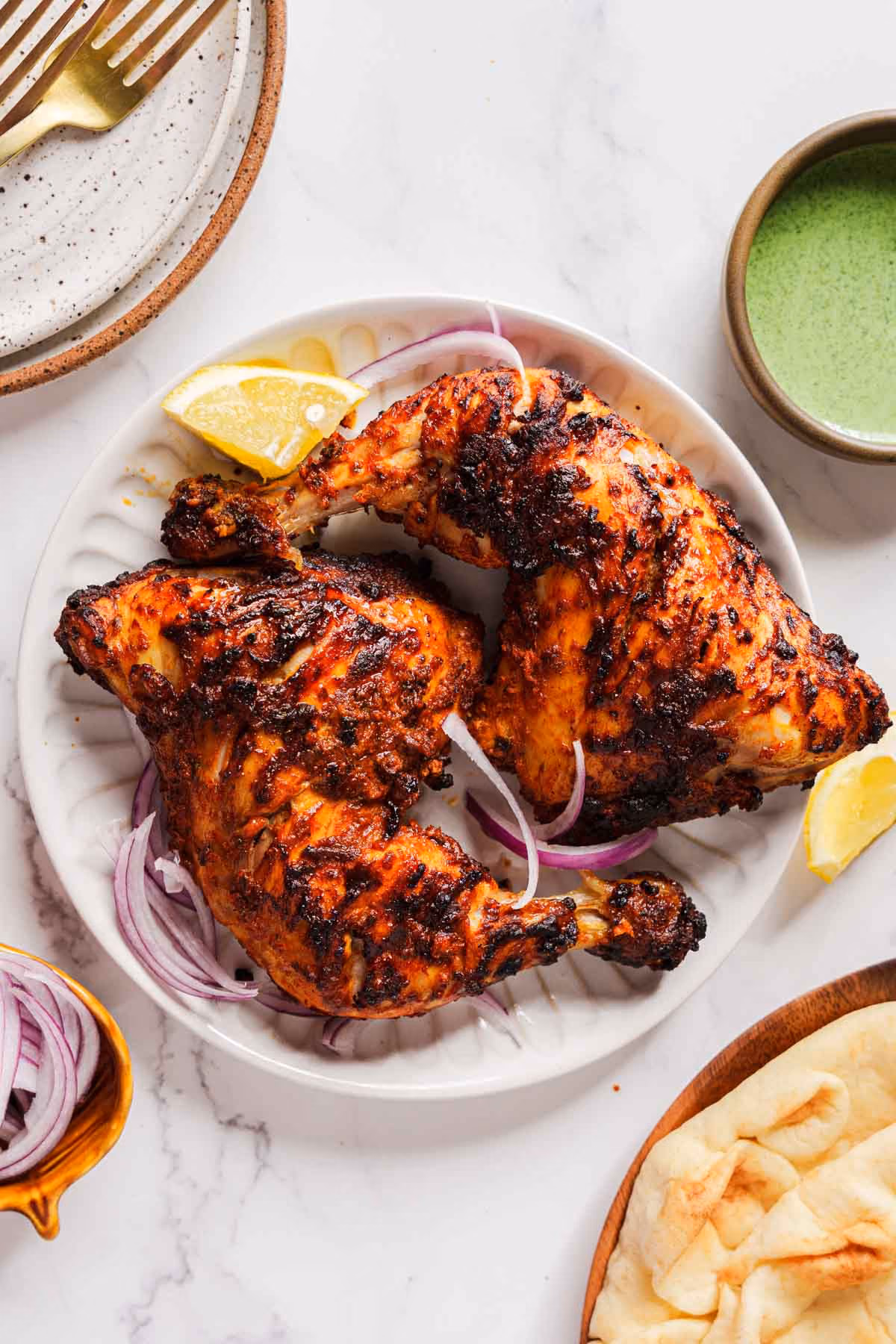 Two pieces of grilled chicken on a white plate with lemon wedges, onions, flatbread, and green sauce.
