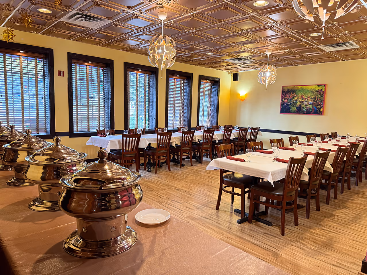 Dining room with long tables set with white tablecloths, glasses, napkins and serving chafers on a side table.